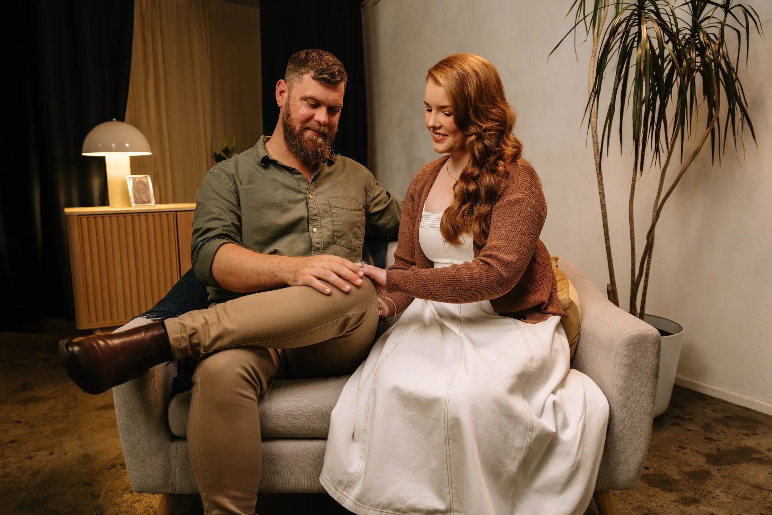 A man and a woman sitting on a beige sofa in a warmly lit living room, sharing an intimate moment with the man holding the woman's hand and she smiling.