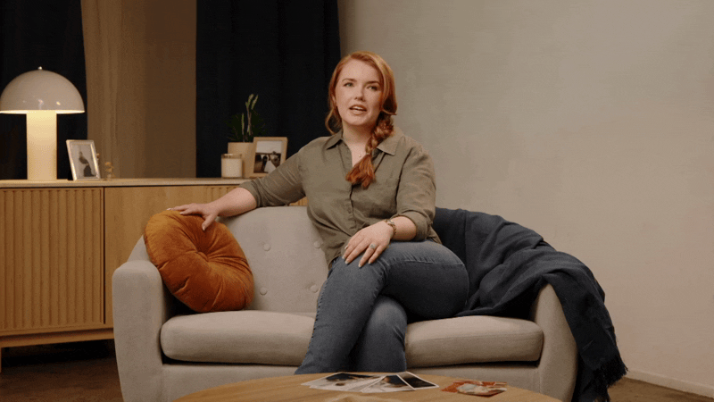 A woman with red hair sitting on a beige couch with a round orange pillow, gesturing while speaking in a living room with a wooden sideboard, framed photos, and a lamp.
