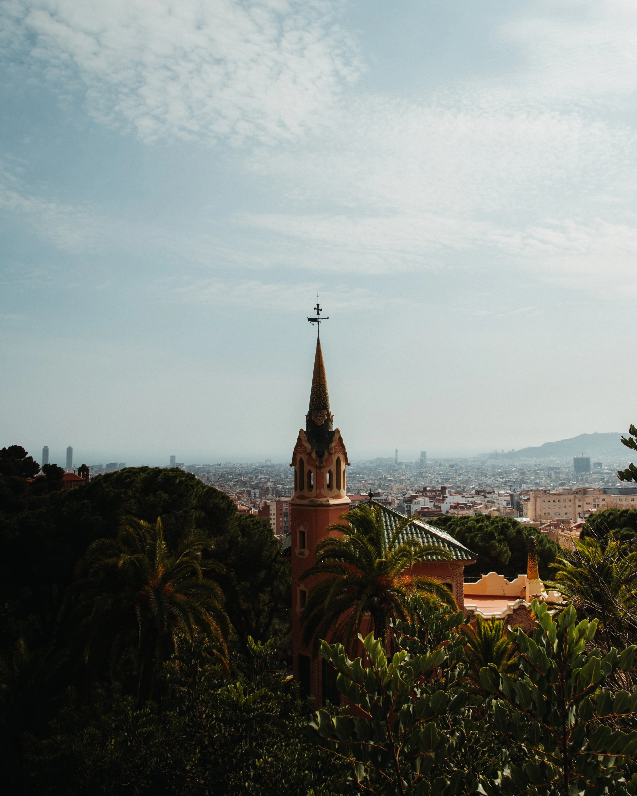 A view of a colorful, ornate building with a tall, pointed steeple, surrounded by lush greenery, overlooking a cityscape under a partly cloudy sky.