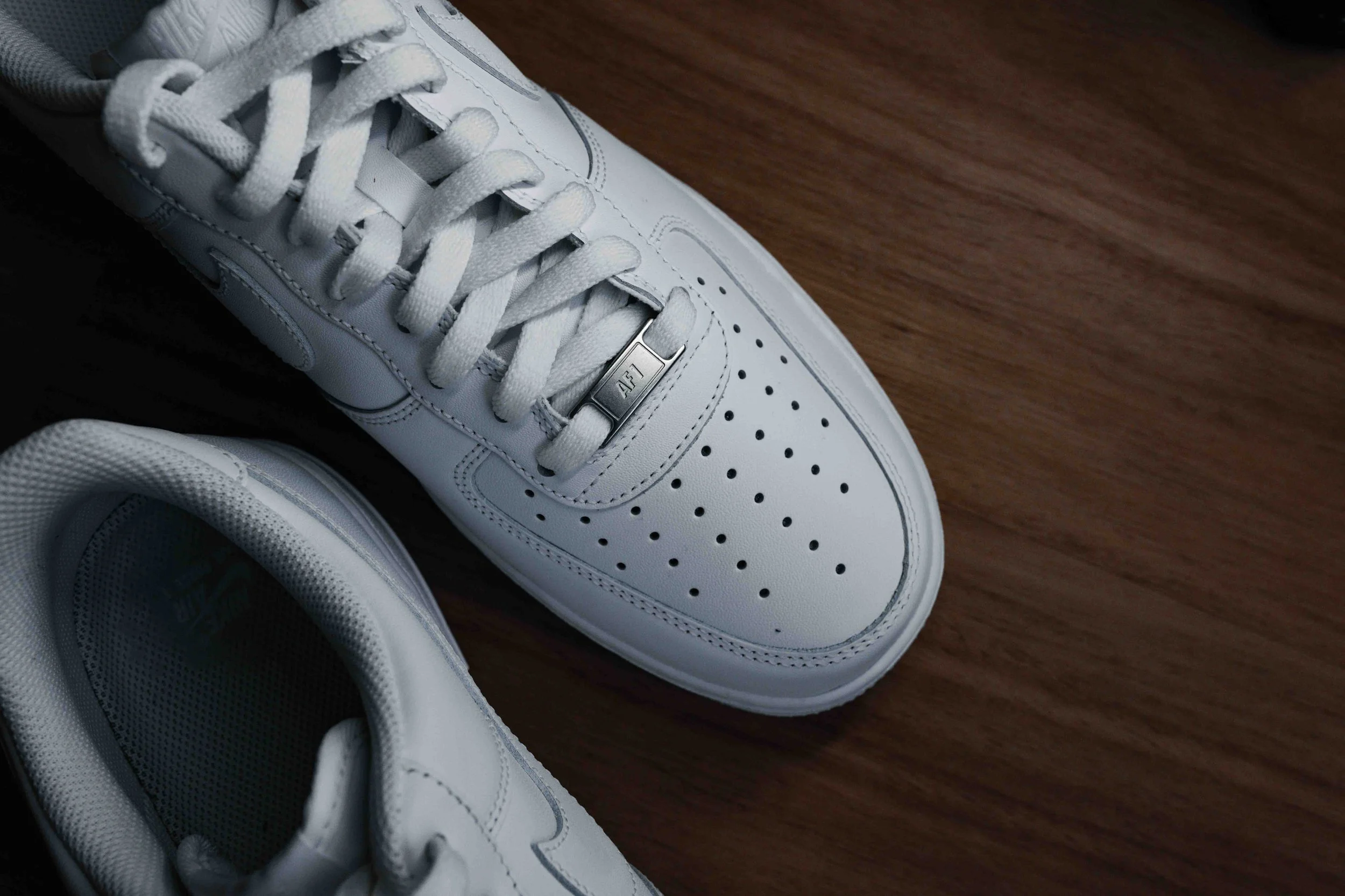 Close-up of a pair of white leather sneakers with laces, metal tag on the laces, and perforated toe box, placed on a wooden floor.
