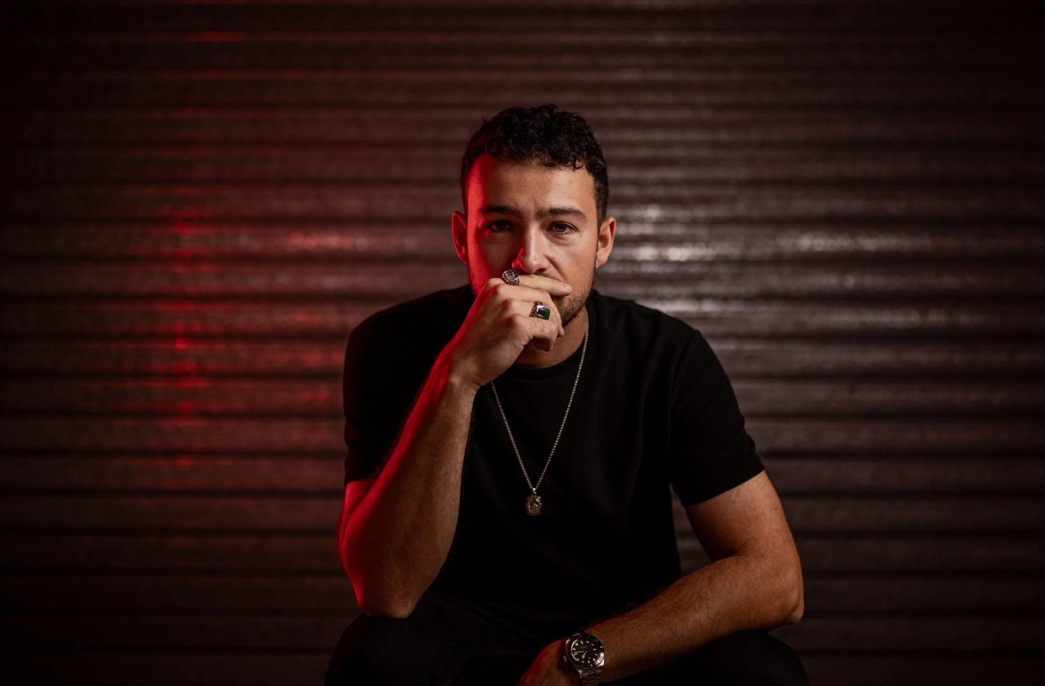 A young man with dark, curly hair sitting in front of a metallic, ridged background. He is wearing a black t-shirt, rings, a necklace, and a watch. He is resting his chin on his hand with a serious expression, with red lighting highlighting one side of his face.