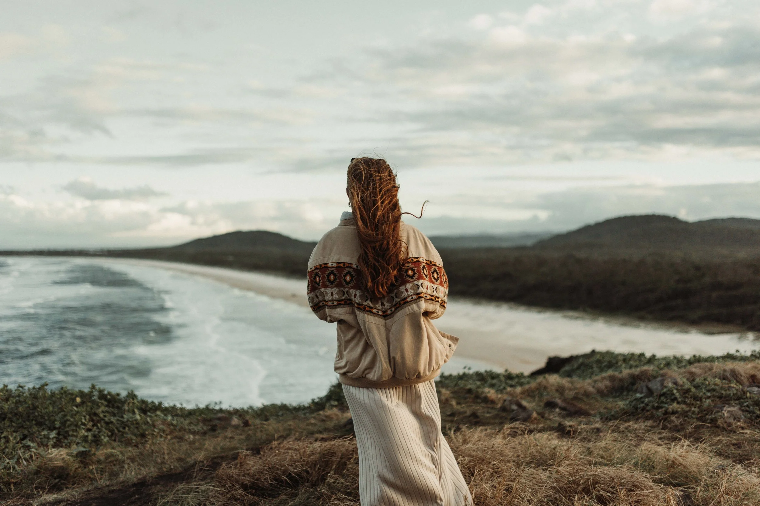 A woman with long, wavy hair standing on a grassy hill overlooking the ocean during cloudy weather, with hills in the background.