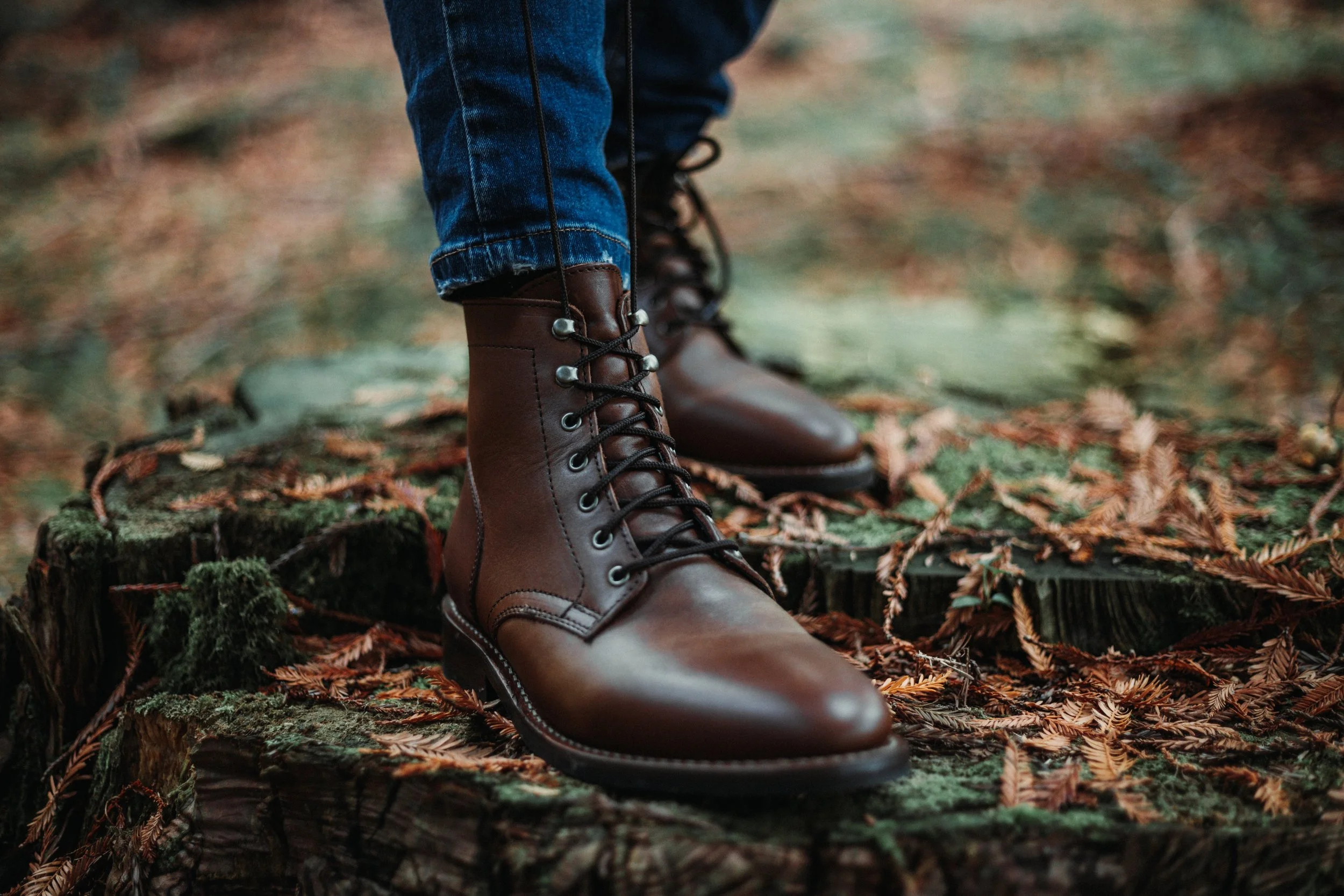 Person wearing brown leather combat boots standing on a mossy, leaf-covered tree stump in a forest with a blurred background.