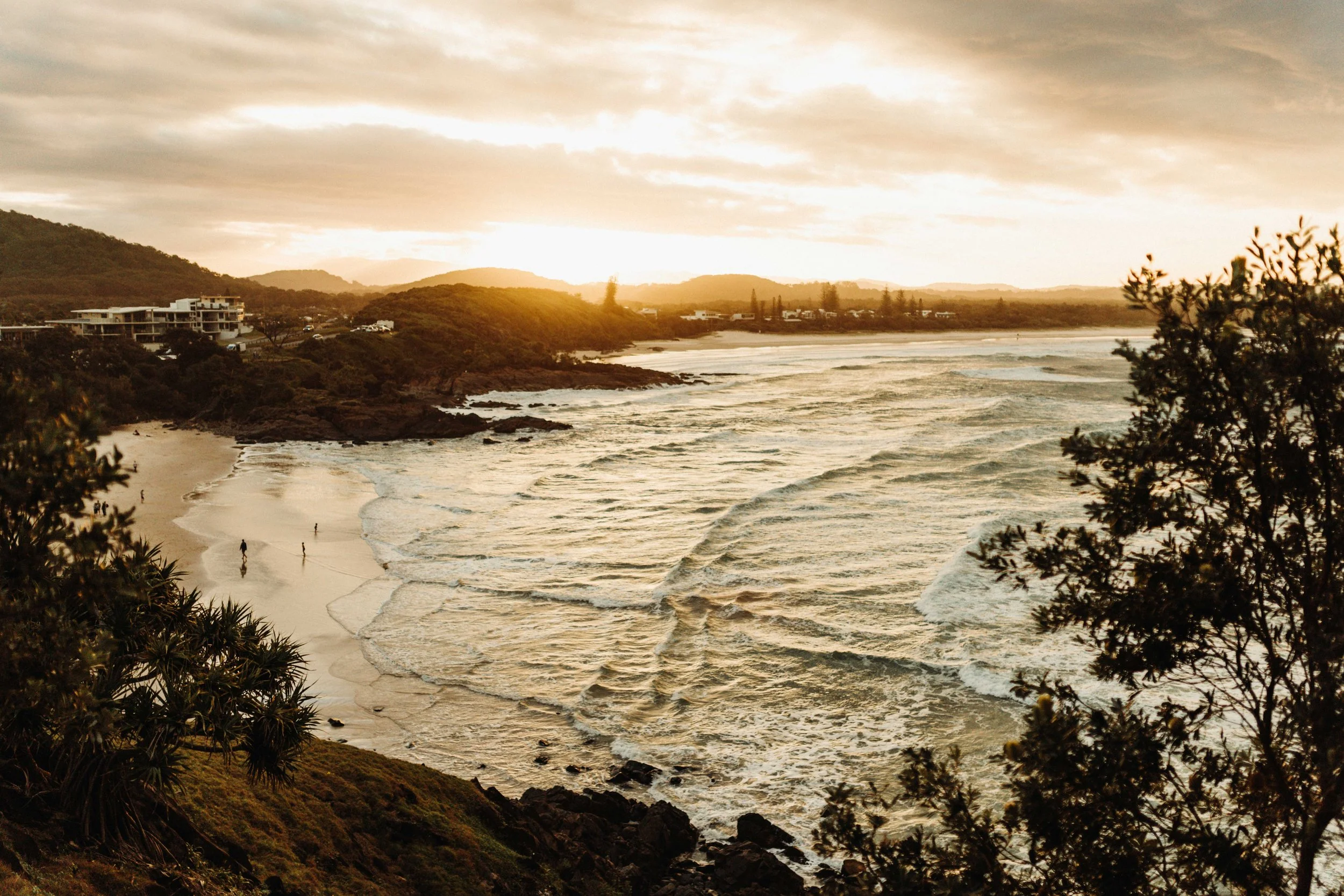 Sunset over a sandy beach with waves, rocks, and foliage in the foreground, and residential buildings on hills in the background.