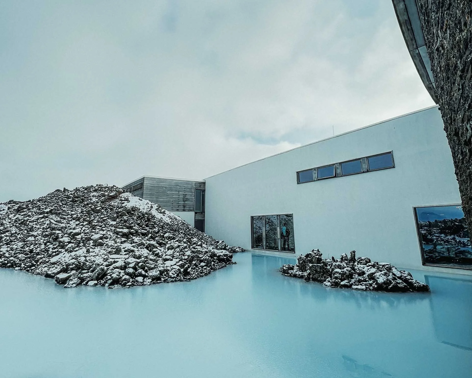 Modern white building with large windows, snow-covered rocks surrounding a frozen blue pond, overcast sky