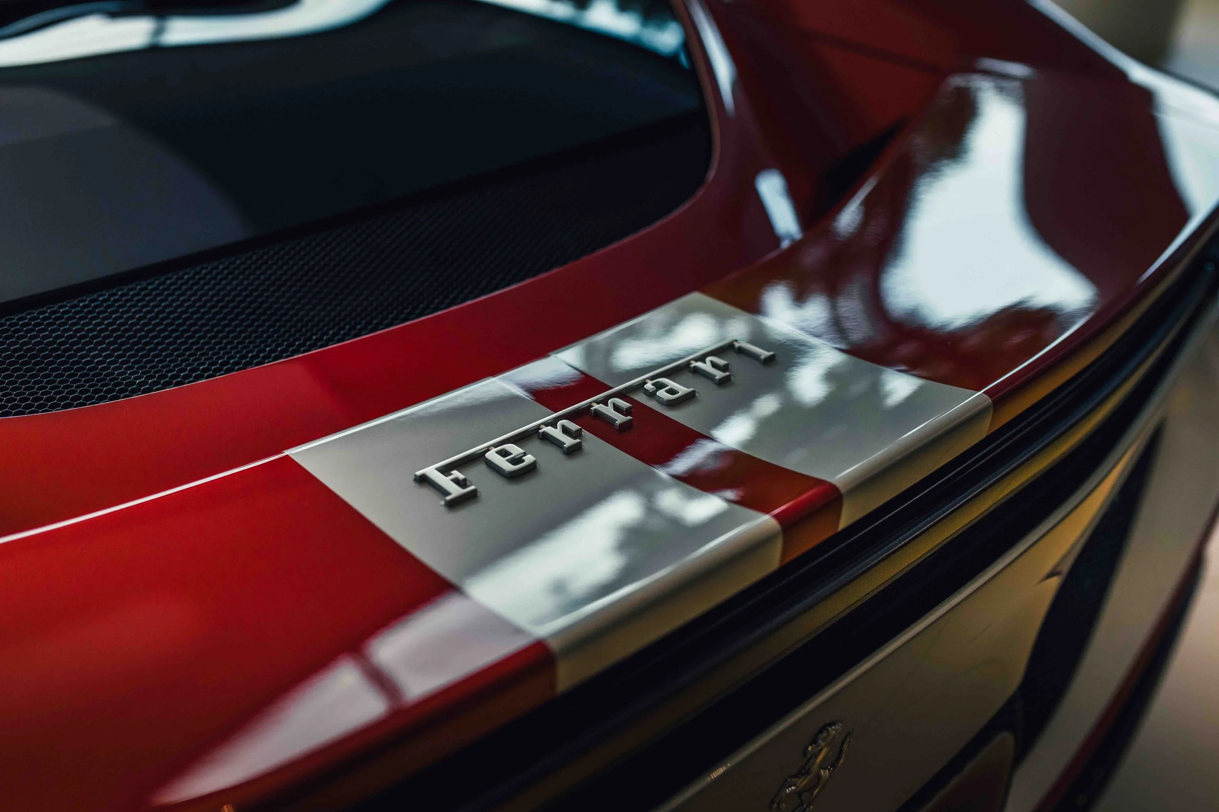 Close-up of a red sports car with a silver Ferrari badge on the rear, reflecting light and surrounding trees.