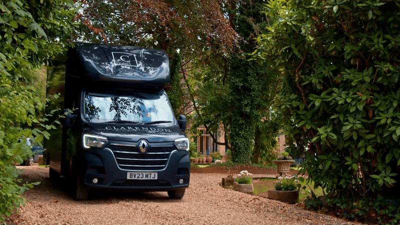 A black delivery van with the name 'Clarenson' on the front, parked on a gravel driveway surrounded by lush greenery and trees.