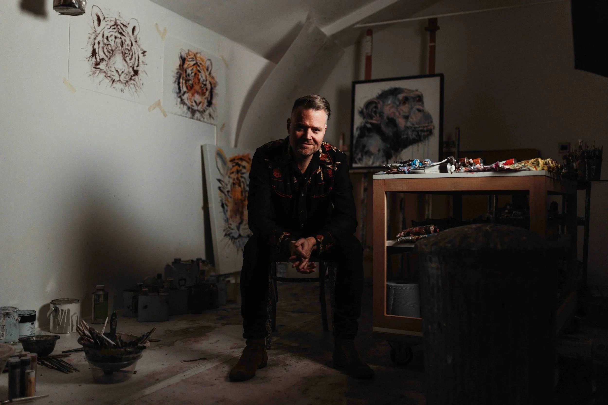 An artist sitting in his studio surrounded by paintings of tigers and a monkey. Art supplies and paintings are visible throughout the room.