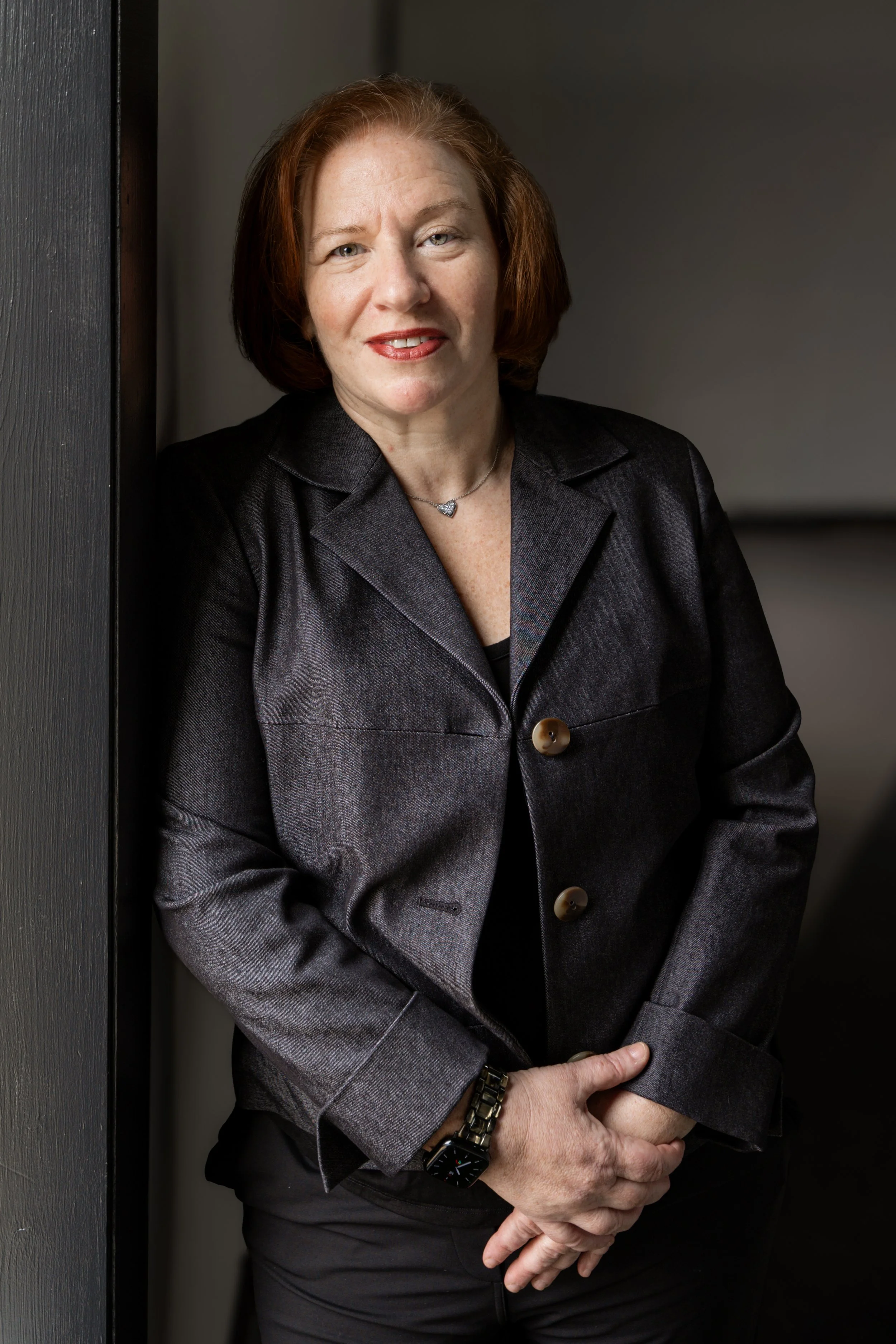 A woman with red hair wearing a dark blazer, standing indoors against a dark wall, looking at the camera with a slight smile.