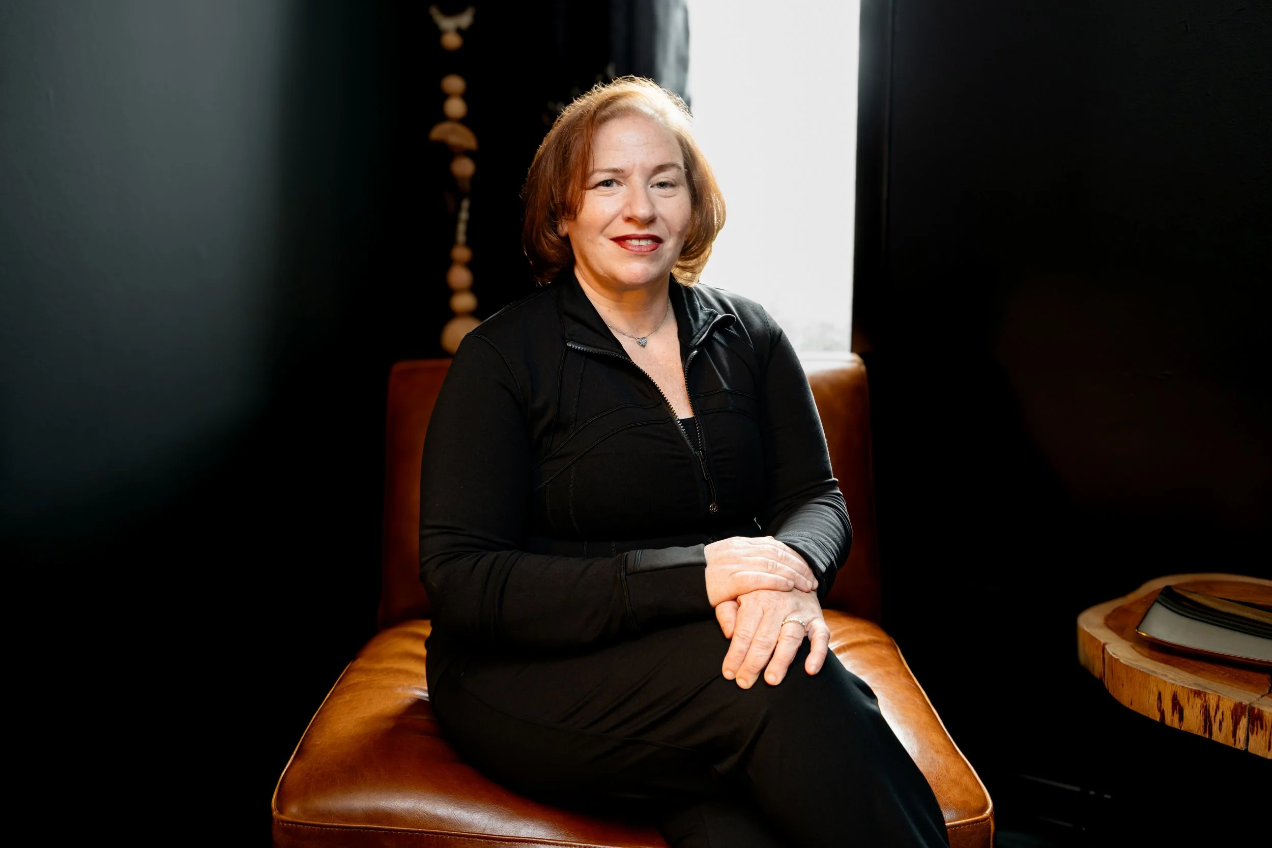 A woman with short red hair wearing a black zip-up jacket and black pants, sitting on a brown leather bench near a window with curtains.