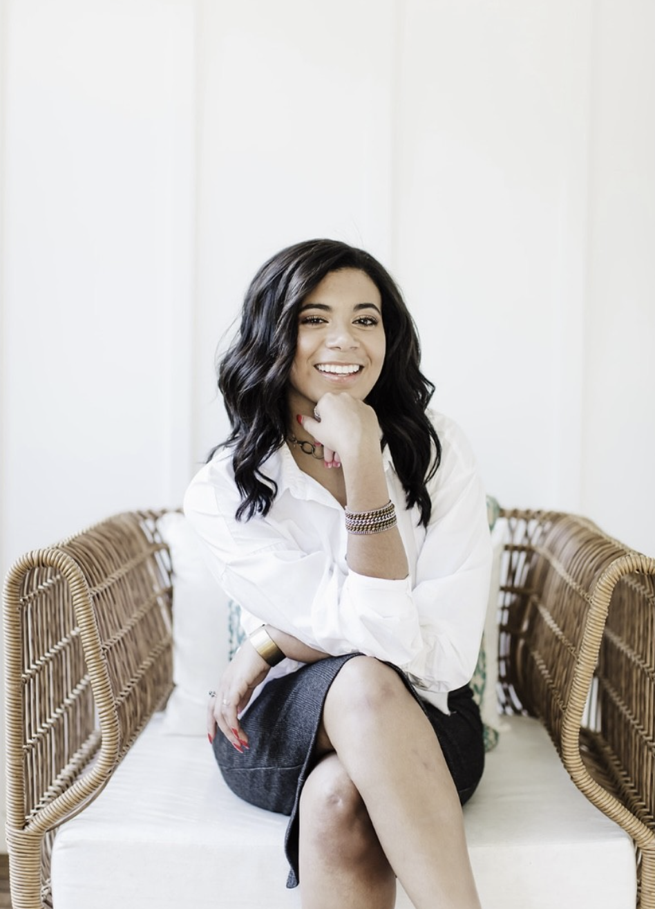 A woman with wavy black hair, wearing a white shirt and dark skirt, sitting on a wicker bench in a bright room, smiling with her hand resting on her chin.