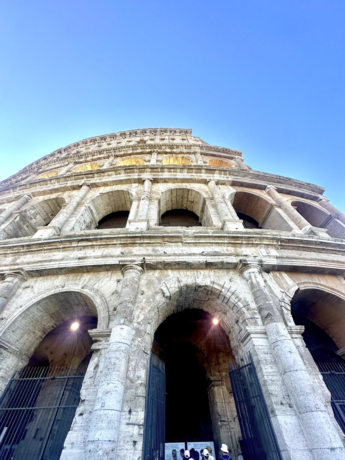 Low-angle view of the Roman Colosseum showing its stone arches and columns against a clear blue sky.