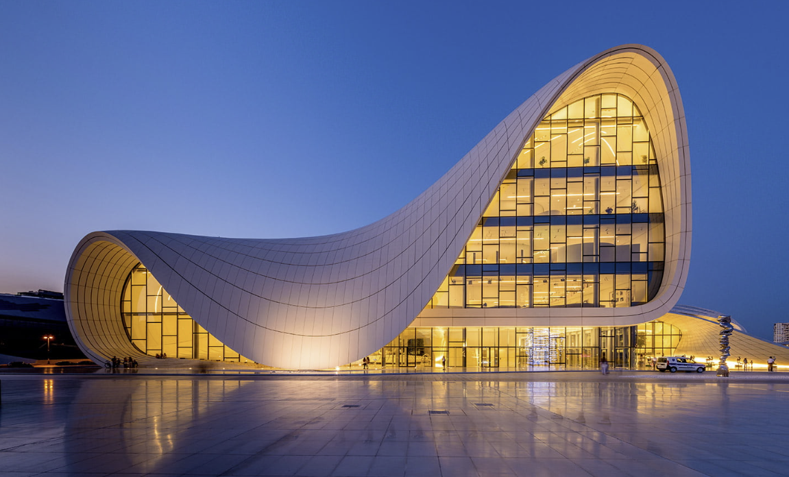 Modern building with curving, wave-like architecture, illuminated from within, at dusk, with a spacious reflective plaza in front.