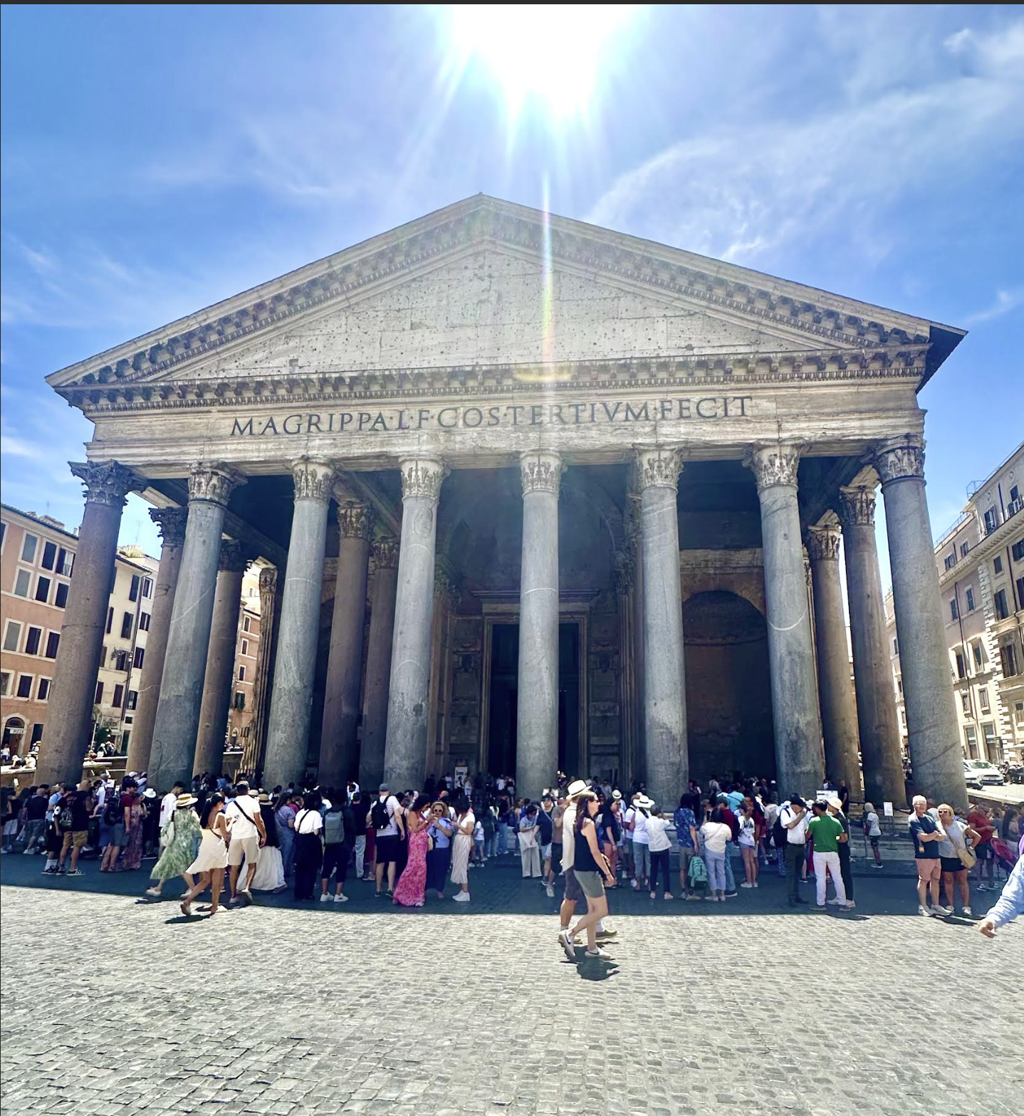 The Pantheon, an ancient Roman temple, with a stone facade and large columns under a bright, sunny sky filled with clouds.