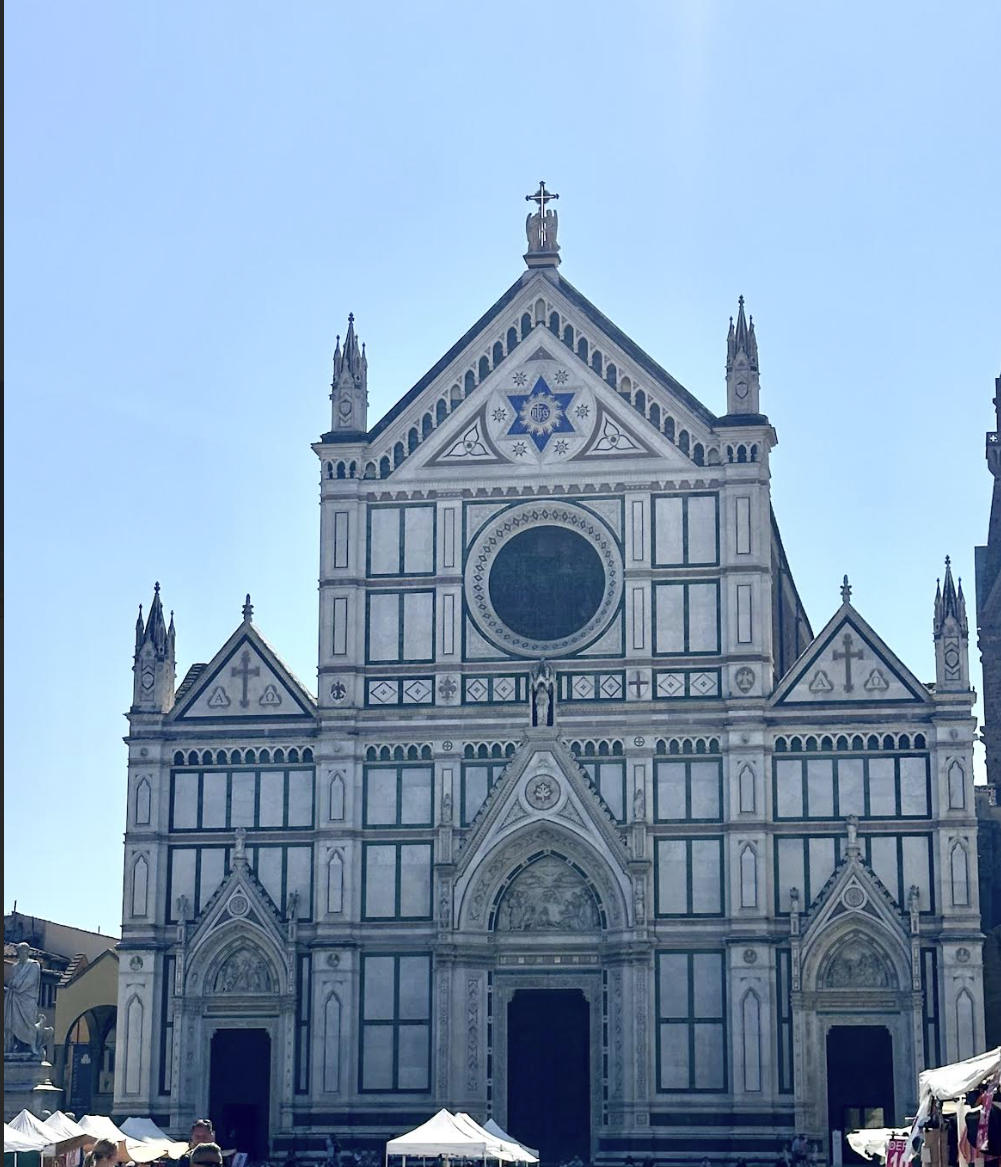 Exterior view of a historic Gothic-style church with white and green marble facade, numerous spires, and intricate architectural details, with market stalls in the foreground.