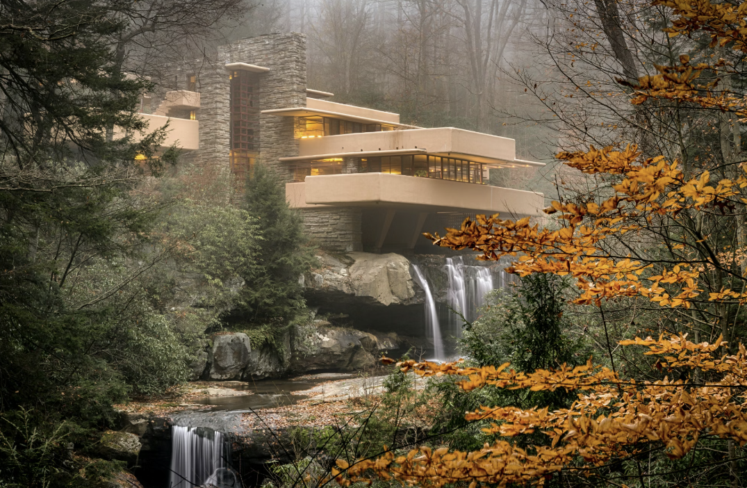 A modern house built on a rocky cliff over a waterfall surrounded by trees with autumn-colored leaves.