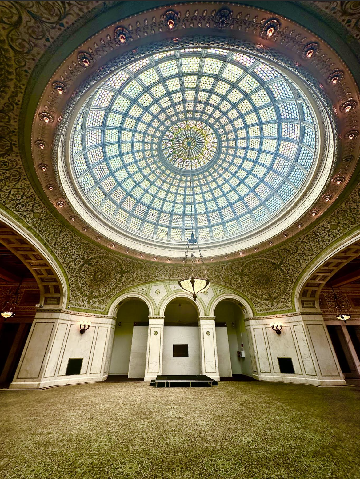 Interior of an elegant room featuring a large stained glass dome ceiling with intricate patterns, decorative moldings, arched doorways, a chandelier, and ornate wall sconces.