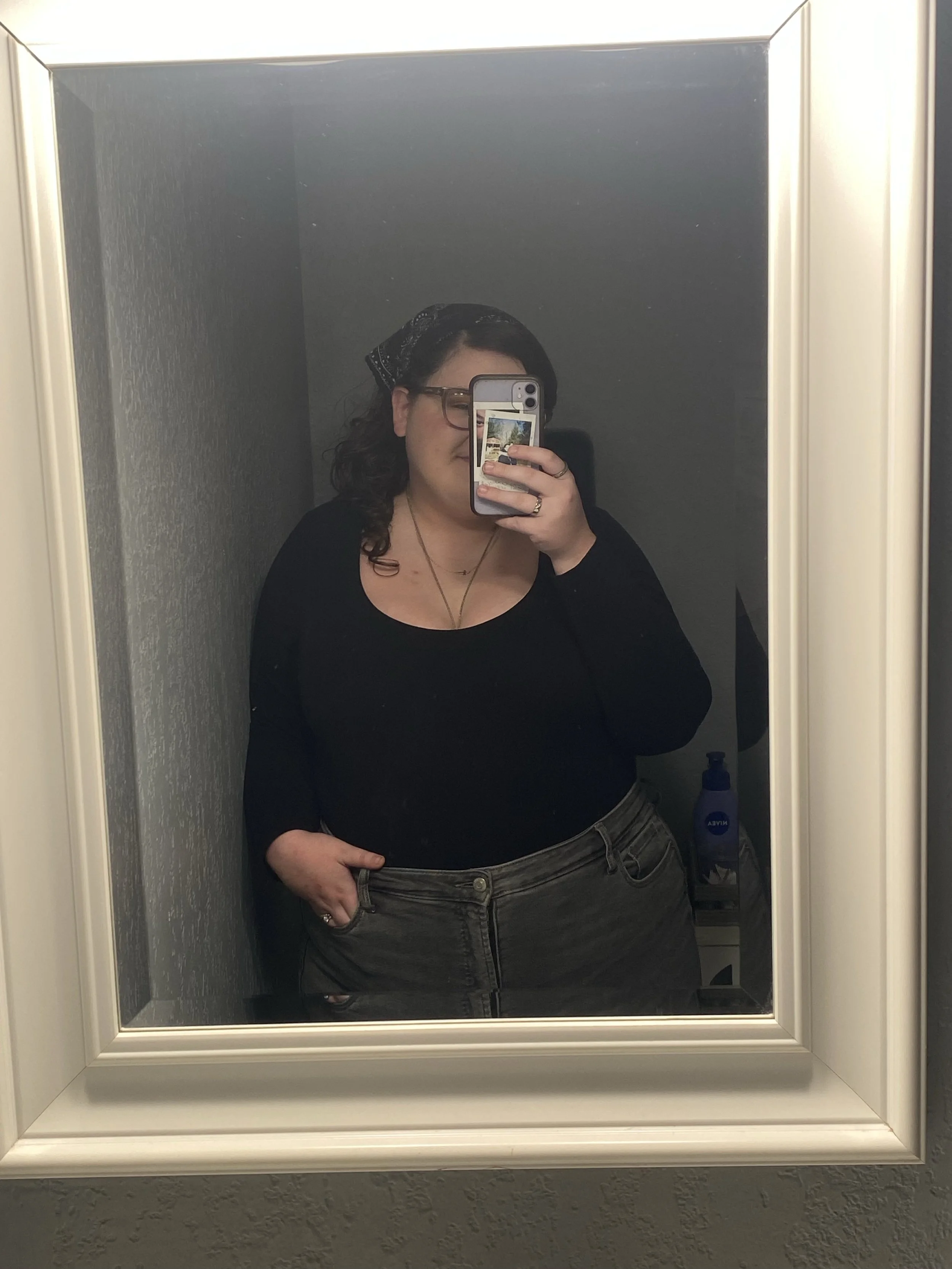 A woman taking a mirror selfie in a bathroom. She is wearing glasses, a black top, and gray jeans. She has dark curly hair, a black bandana, and is holding a phone with a colorful case.