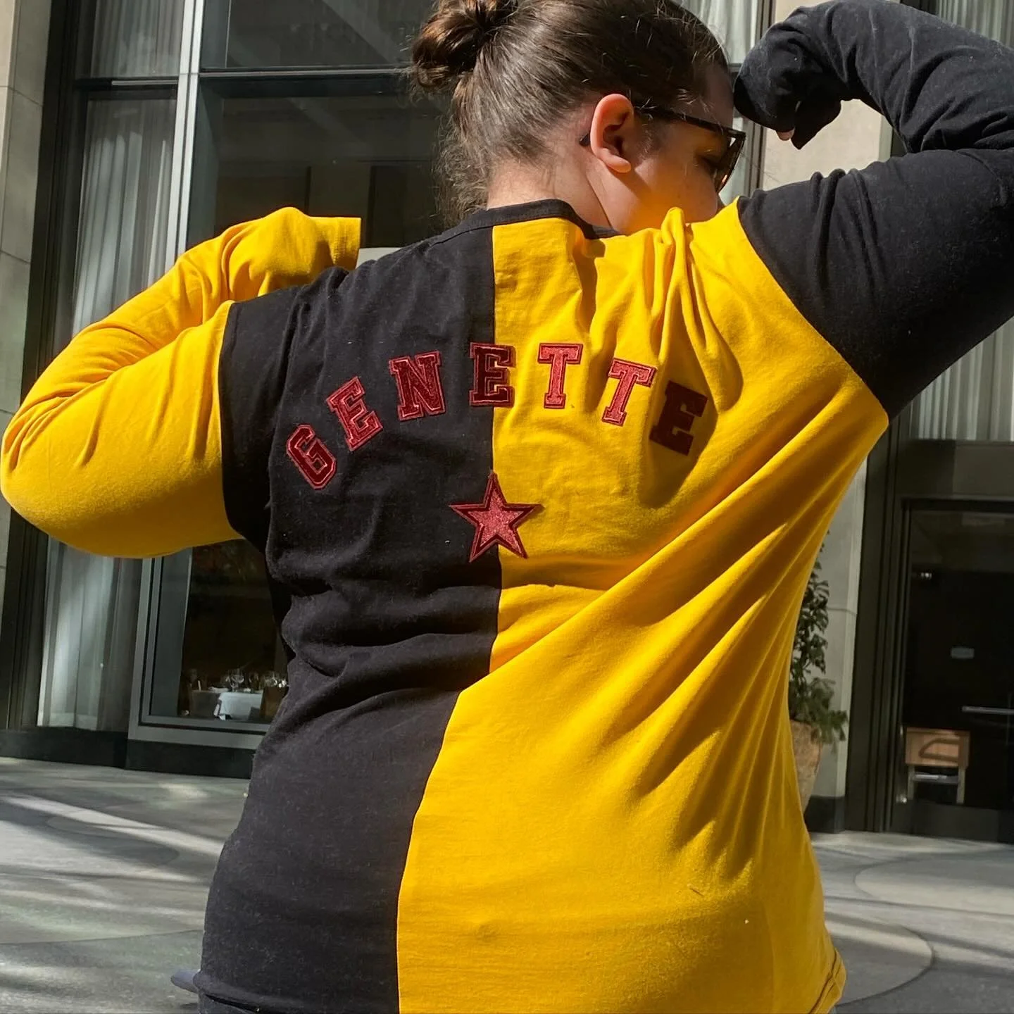 A person with brown hair in a bun wearing sunglasses and a black and yellow varsity jacket with 'GENETTE' embroidered on the back and a star emblem, outdoors near a building with glass doors.