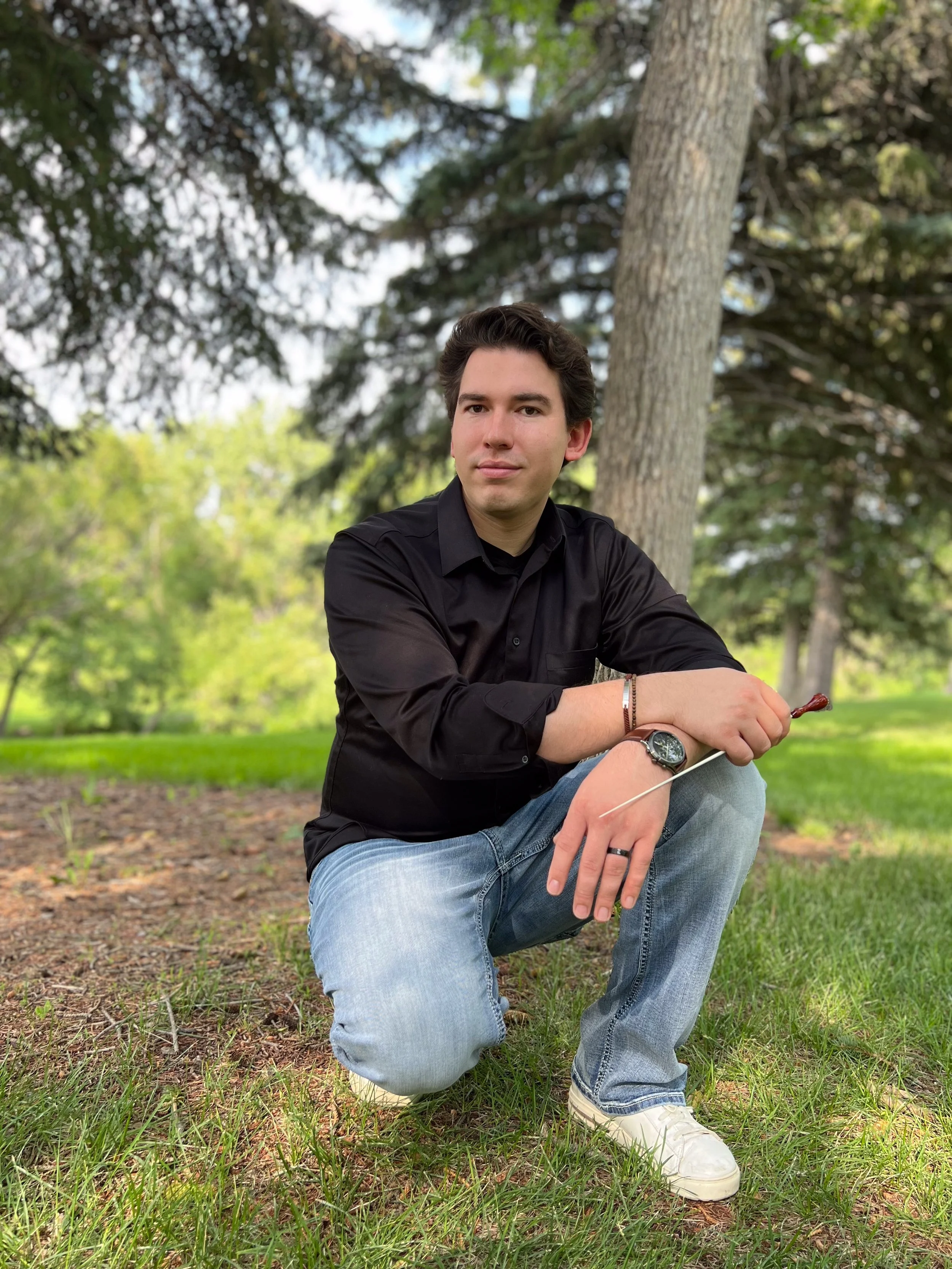 A young man with dark hair, wearing a black shirt and light blue jeans, is kneeling on one knee on a grassy area outdoors, holding a conductor's baton, with trees and green foliage in the background.