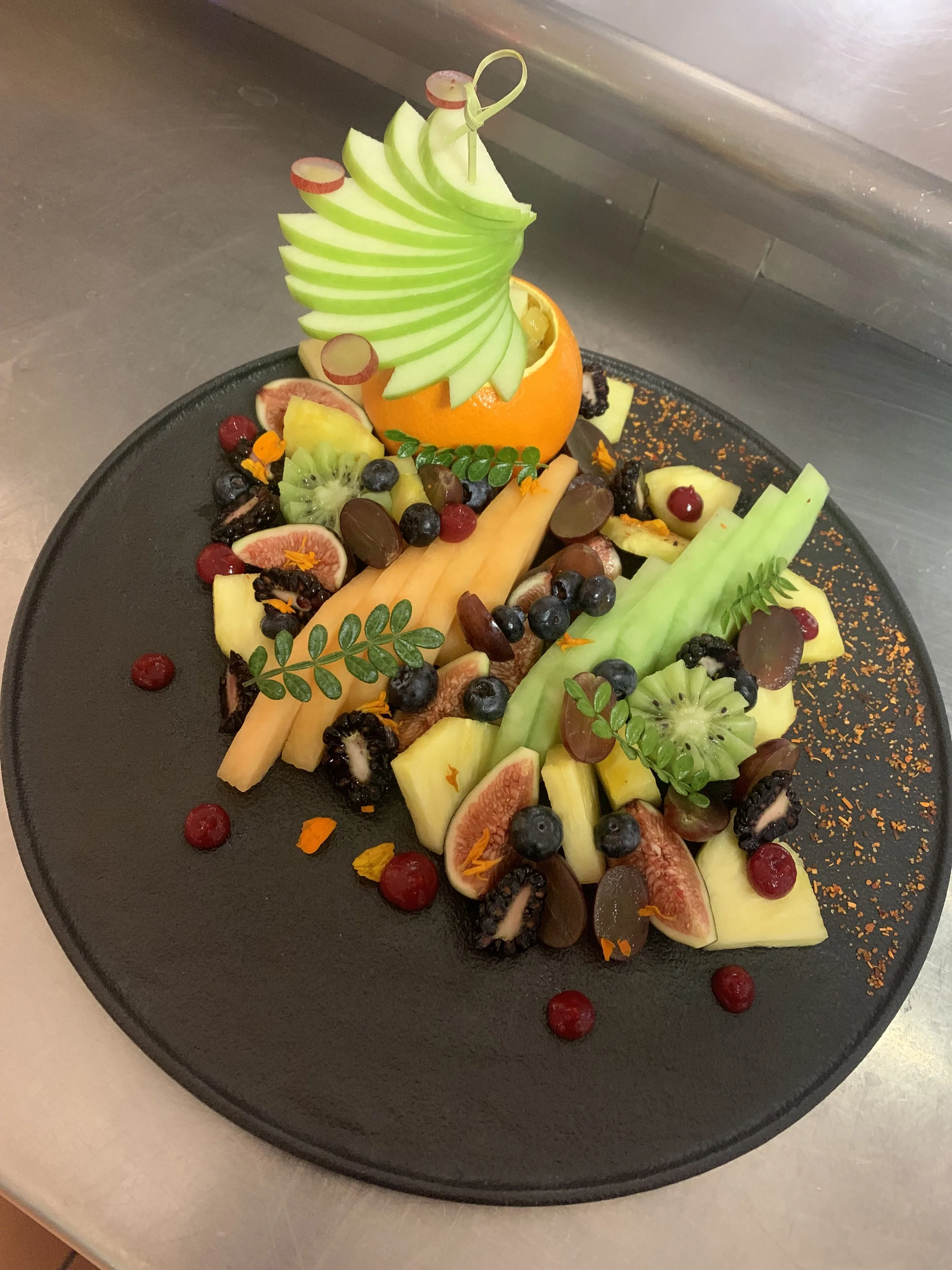 A fruit platter arranged creatively on a black plate, featuring slices of kiwi, fig, cantaloupe, honeydew, grapes, and orange, with decorative garnishes and edible flowers.