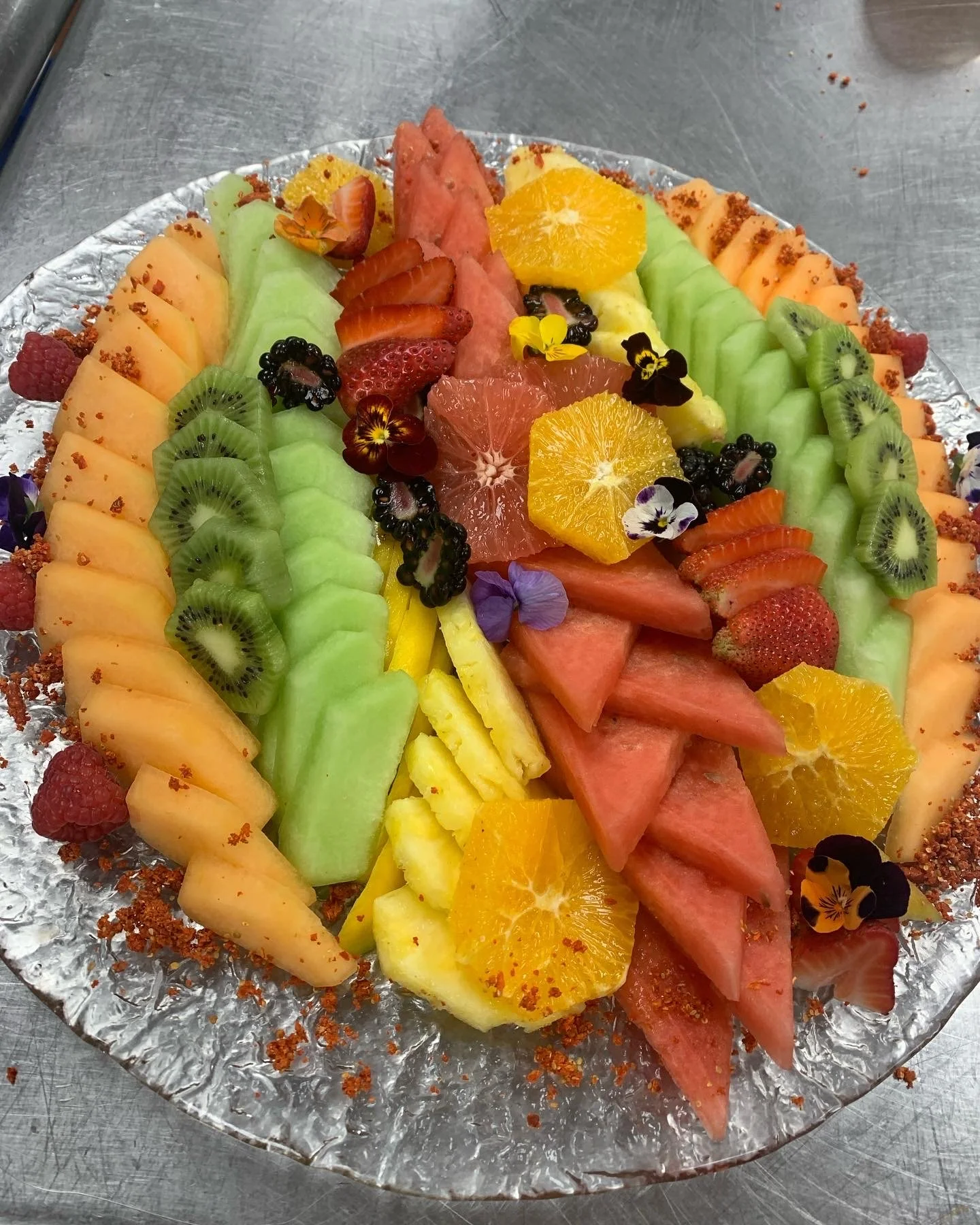 A colorful fruit arrangement with slices of watermelon, cantaloupe, honeydew, orange, strawberry, kiwi, and blackberries, garnished with edible flowers on a silver foil tray.