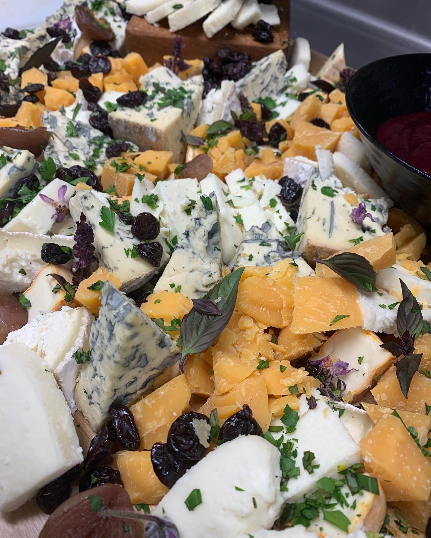 A cheese platter with a variety of cheeses, dried raisins, and herbs, garnished with fresh herbs and edible flowers.