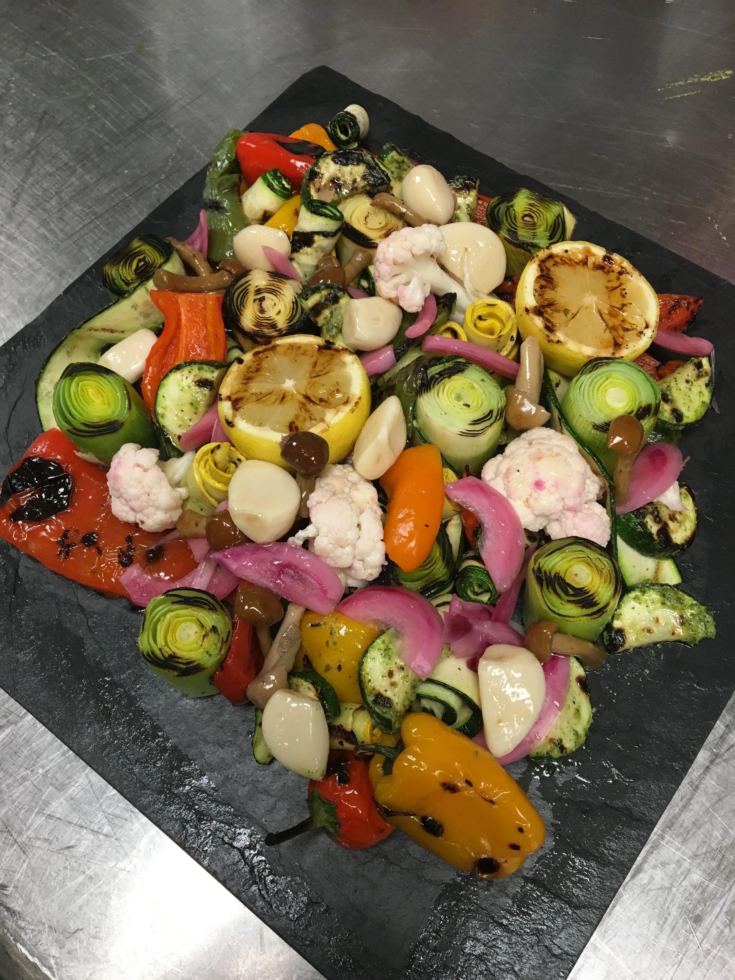 Grilled vegetable platter on black slate, including grilled lemon halves, grilled zucchini, red and yellow bell peppers, pickled red onions, and assorted mushrooms.