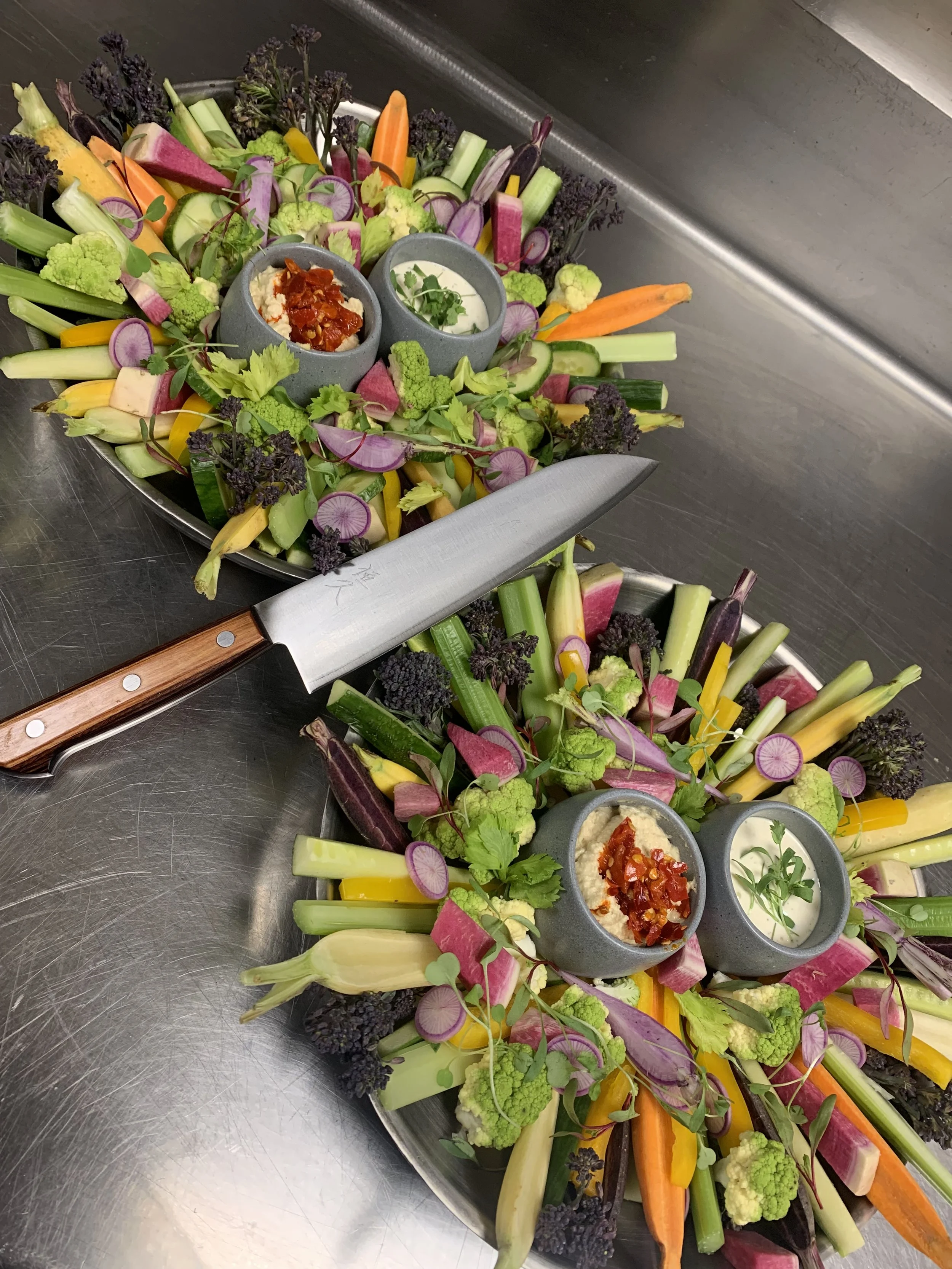 Colorful vegetable platter with dipping sauces in gray bowls, including hummus topped with spicy peppers and a creamy sauce garnished with herbs, surrounded by sliced carrots, cucumbers, radishes, celery, cauliflower, and purple lettuce on a metal tr