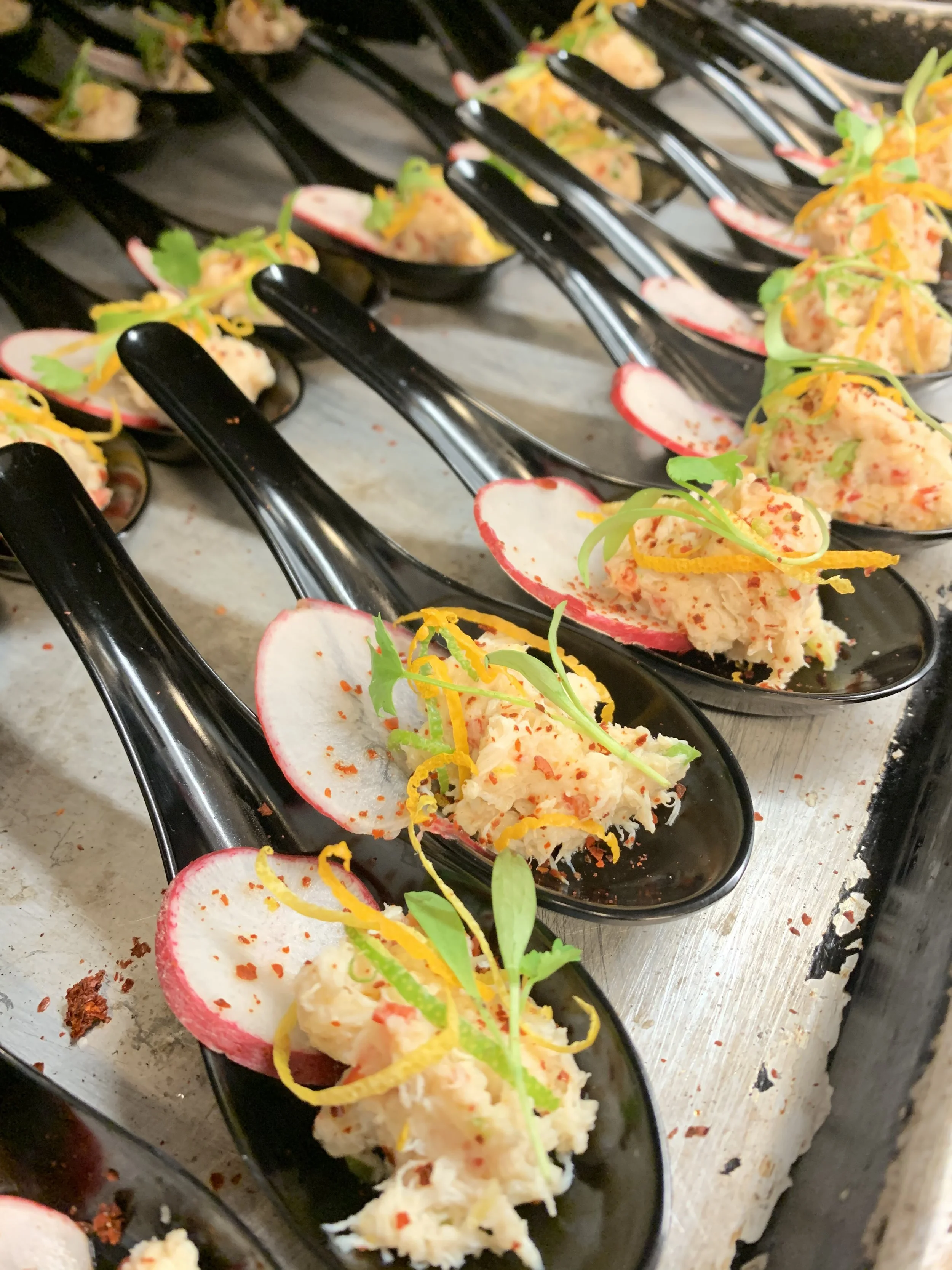 Close-up view of small black dishes of potato salad garnished with radish slices, shredded cheese, microgreens, and red pepper flakes on a metal countertop.