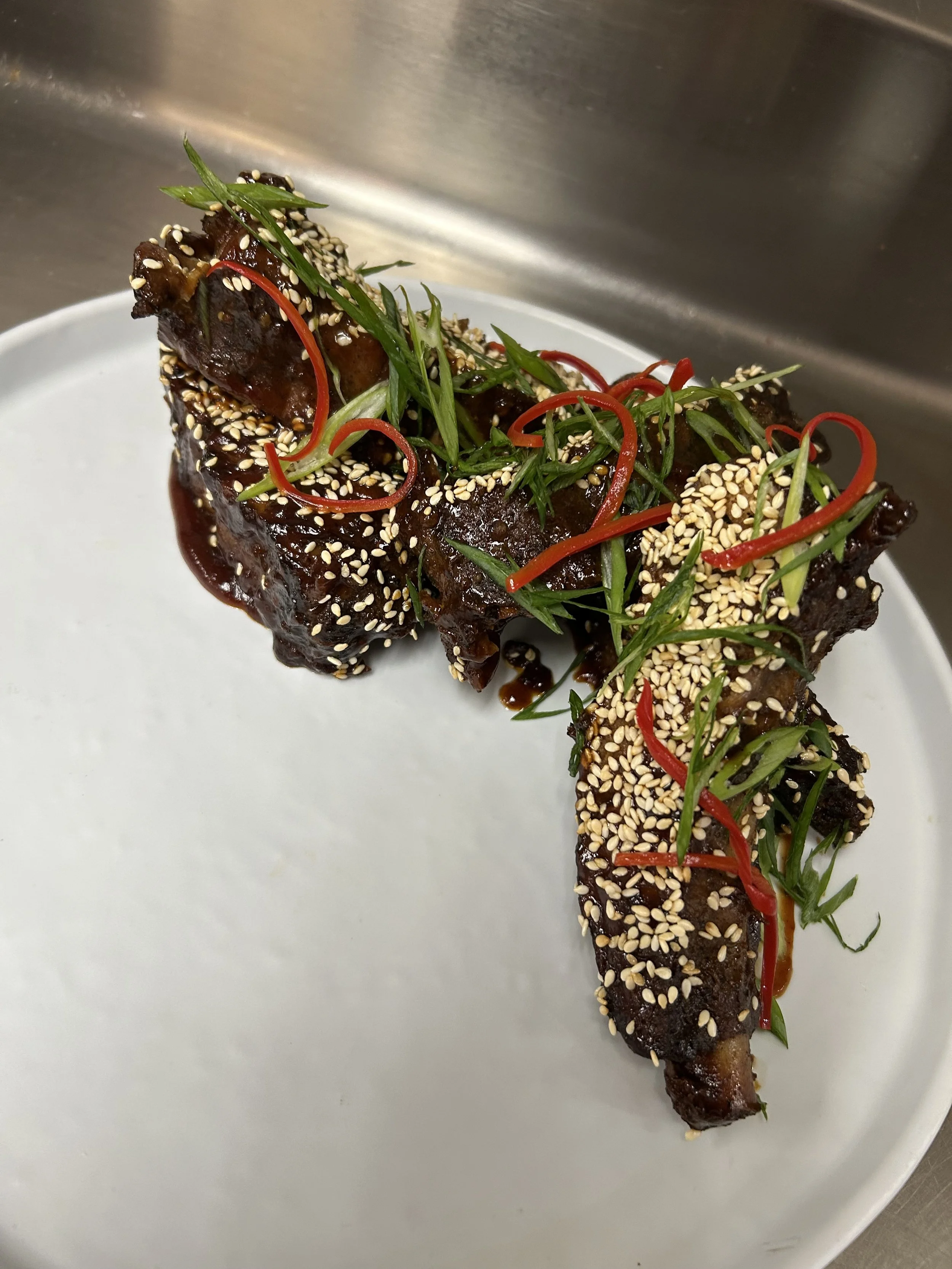 A dish featuring a piece of glazed, dark-colored meat garnished with sesame seeds, sliced red chili peppers, and chopped green herbs or scallions, served on a white plate.