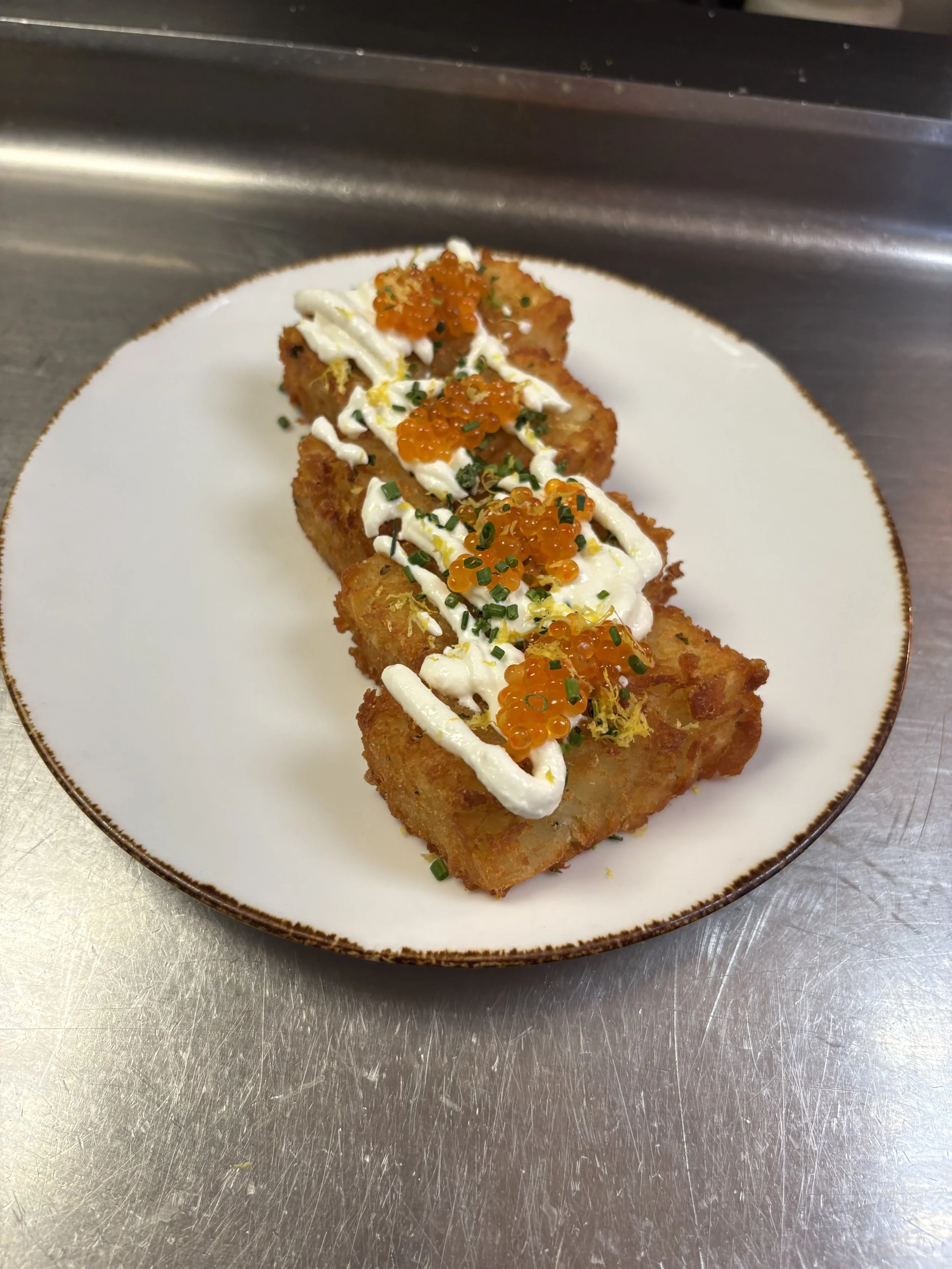 Fried dish topped with white sauce, orange fish roe, and chopped chives on a white plate with a brown rim, placed on a stainless steel surface.