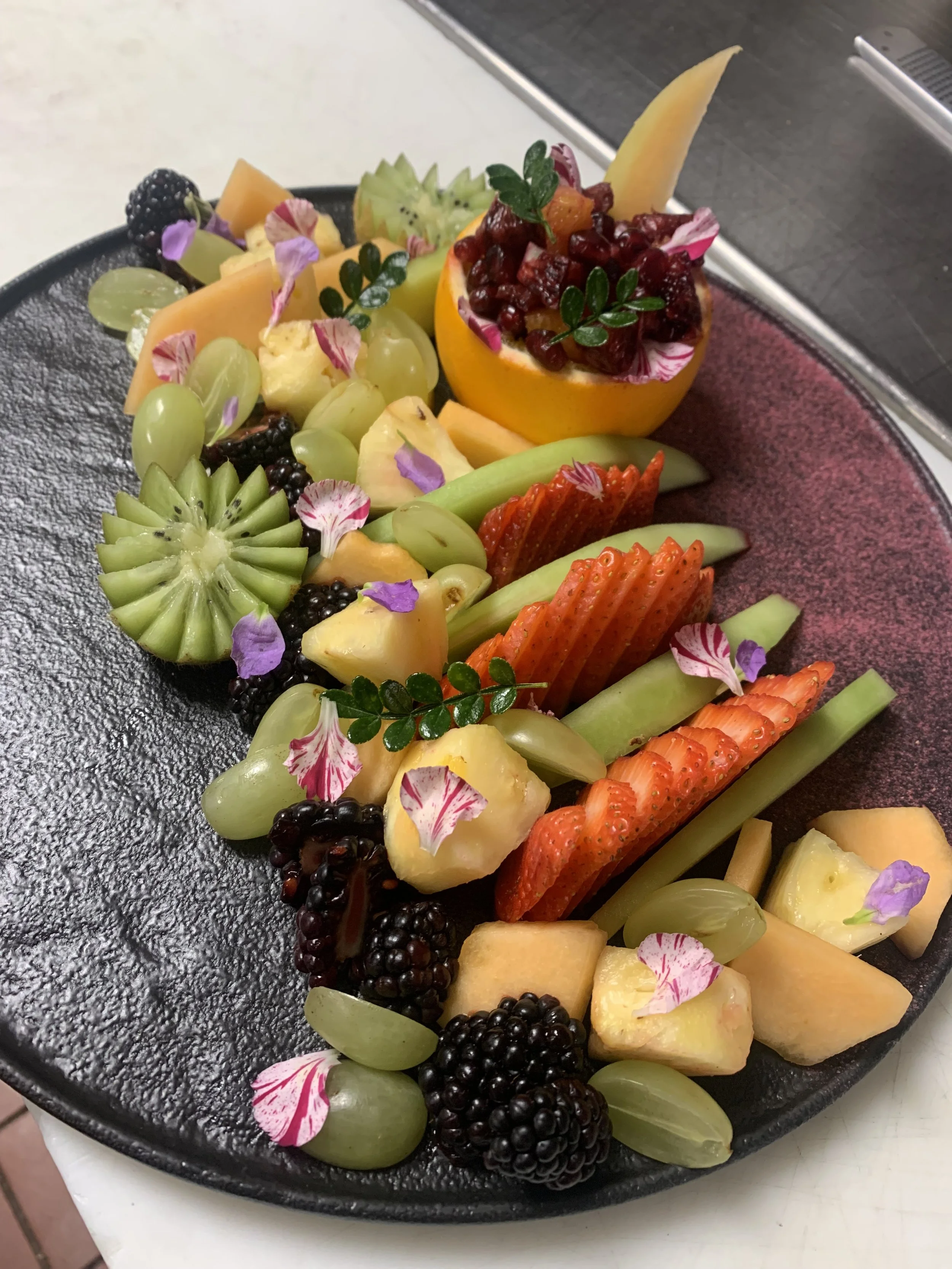 Assorted fresh cut fruits arranged on a black plate, garnished with edible flowers and small green leaves.