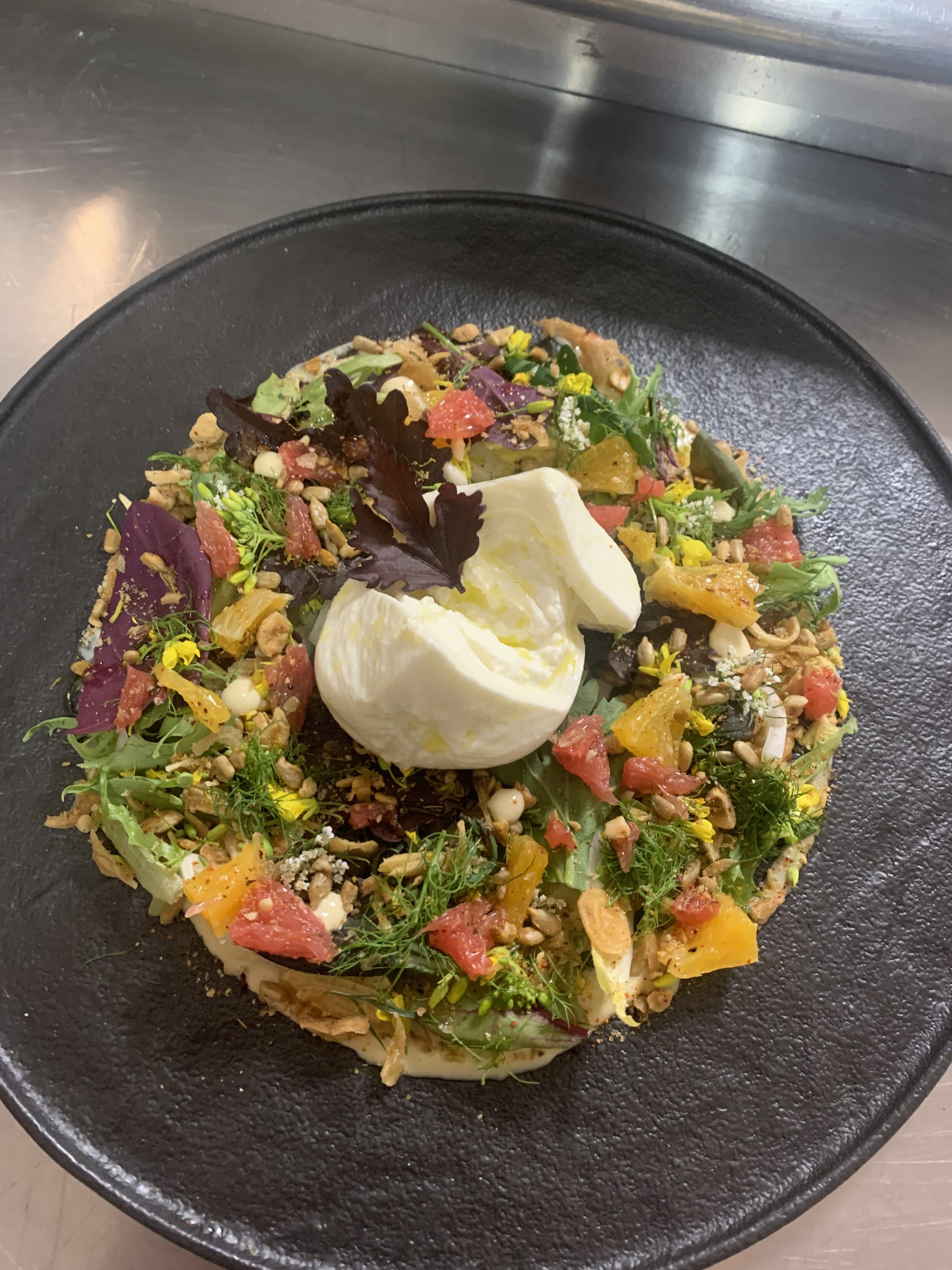 A plated salad on a black round plate, featuring mixed greens, colorful diced fruits like grapefruits, soft cheese, seeds, herbs, and edible flowers.