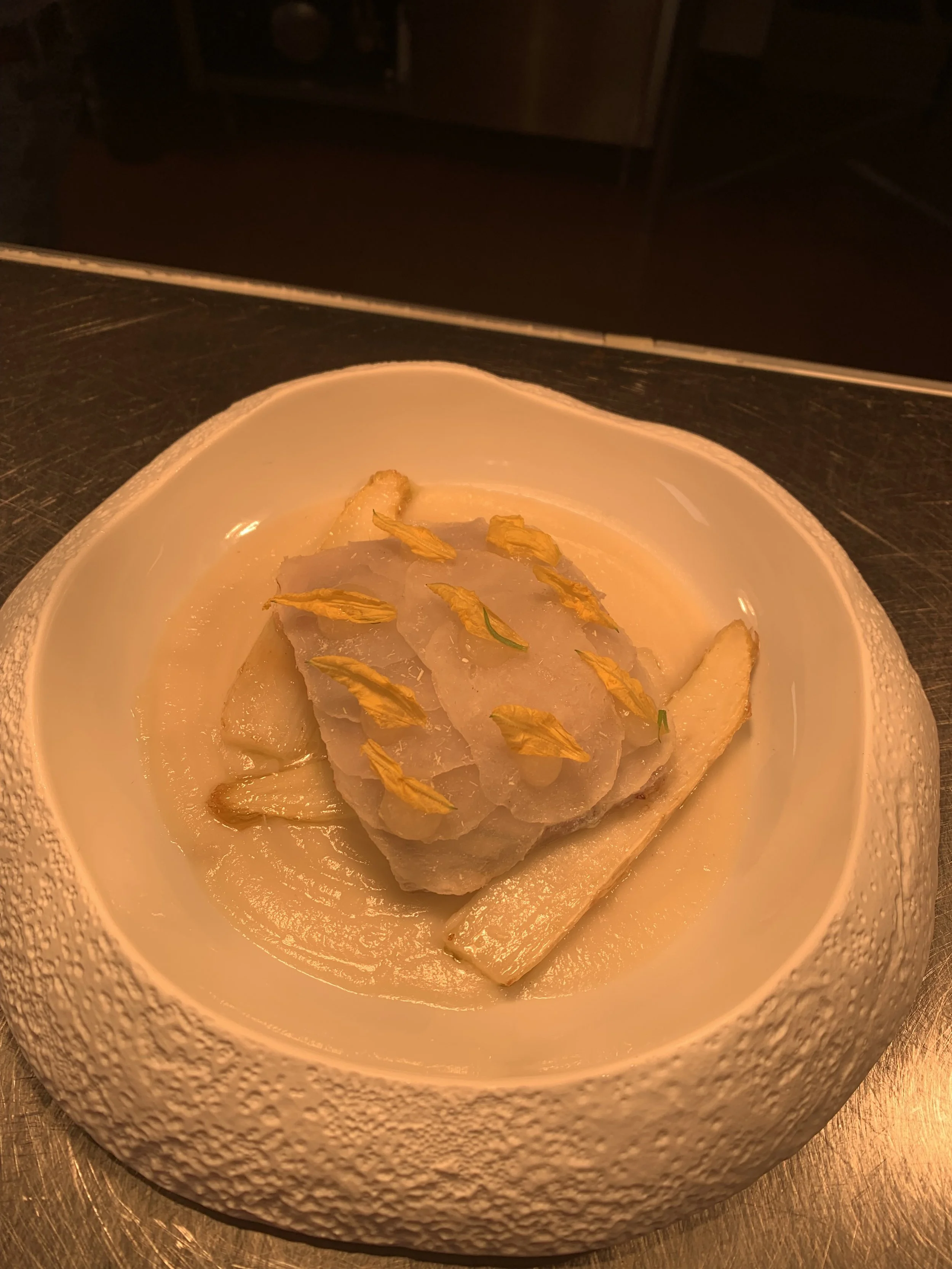 A white textured bowl containing a cooked fish fillet garnished with small edible yellow flowers, served on a wooden surface.