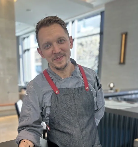 A man in a gray chef's coat and apron standing indoors with large windows in the background, smiling at the camera.