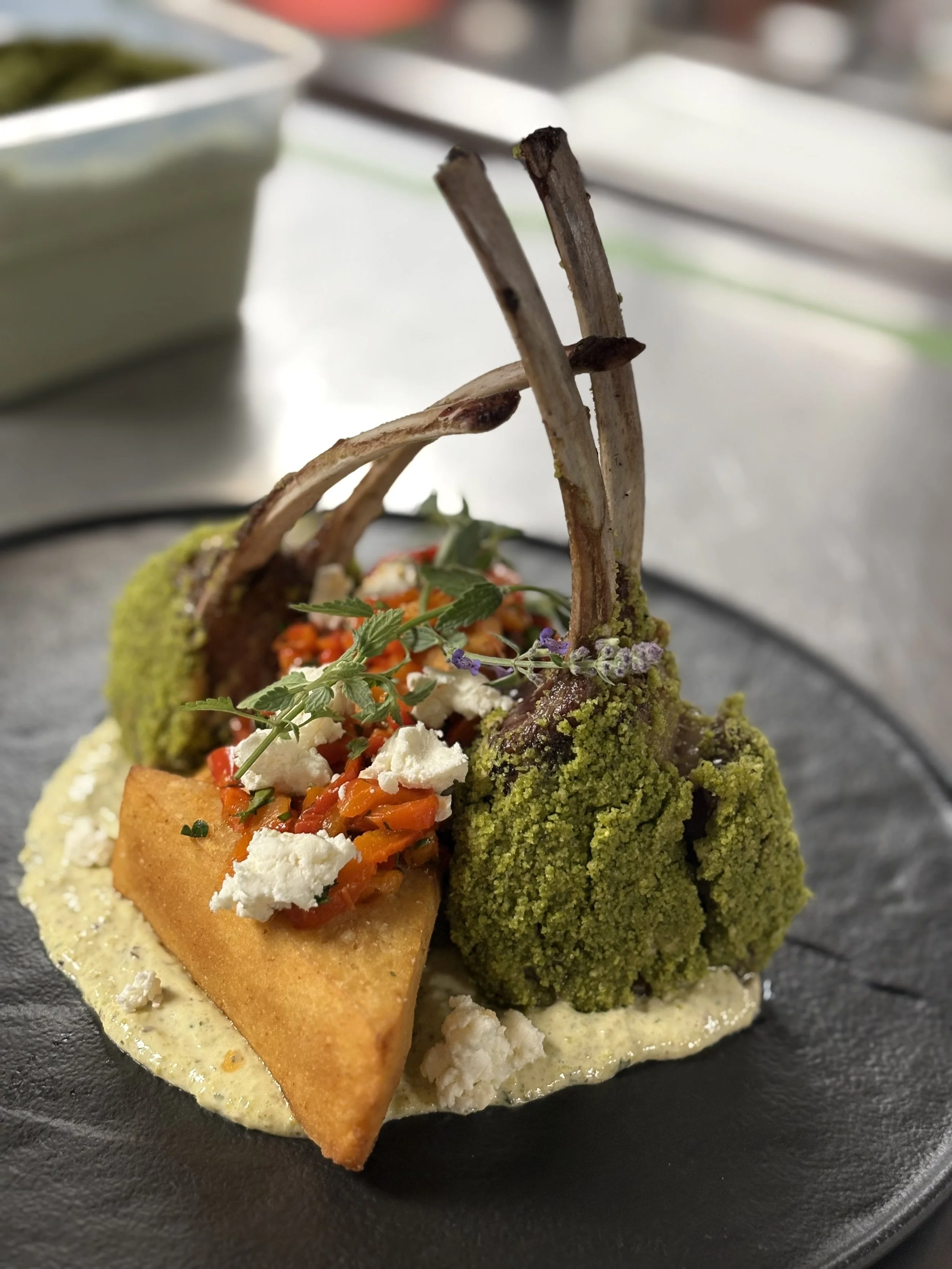 Close-up of a gourmet dish featuring breaded protein, green and purple vegetables, and garnishes, served on a dark plate.
