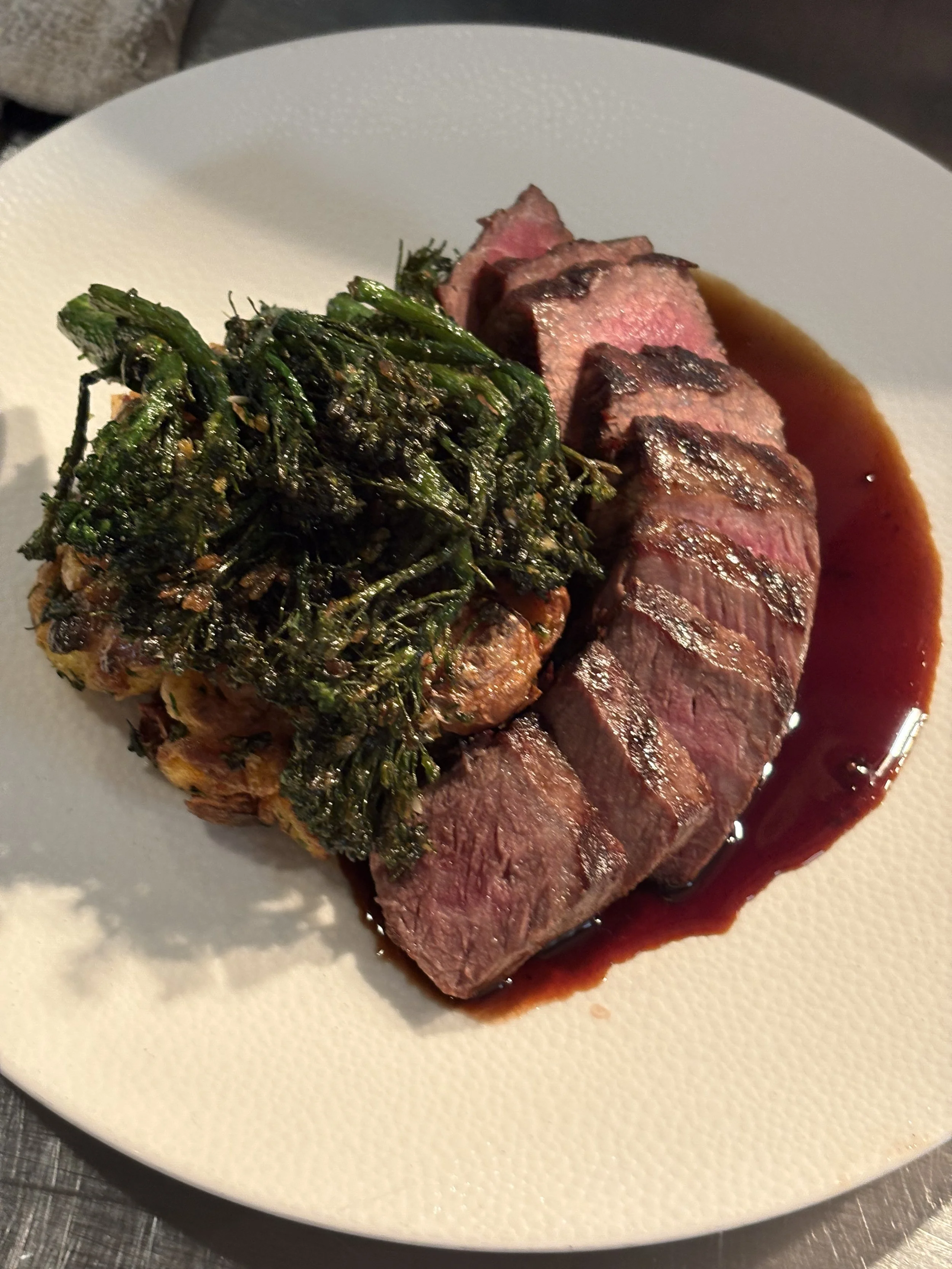 Slices of medium-rare steak with a dark sauce, topped with a portion of cooked greens on a white textured plate.