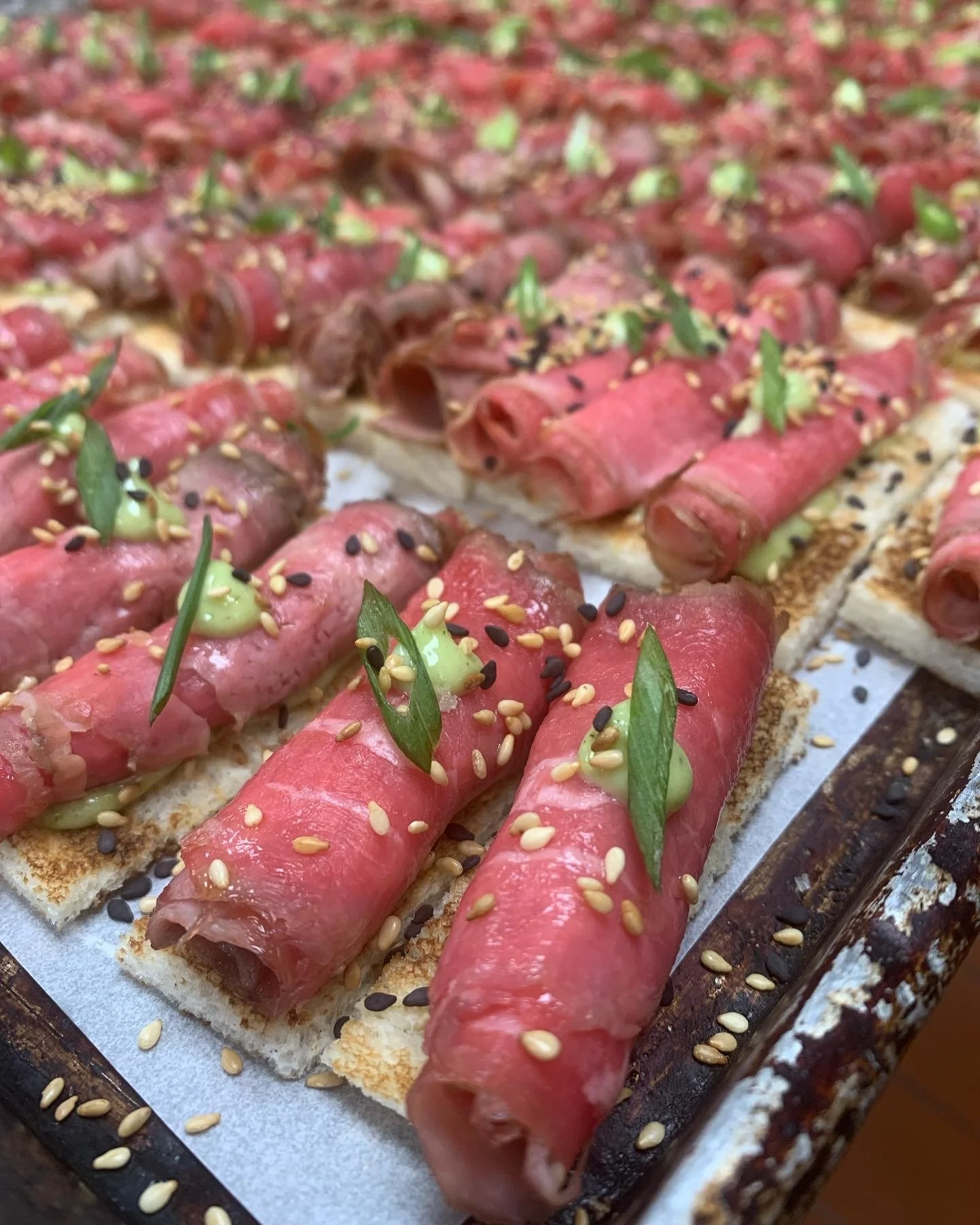 Close-up of bite-sized appetizers with slices of meat, green sauce, and sprinkled sesame seeds on a tray.