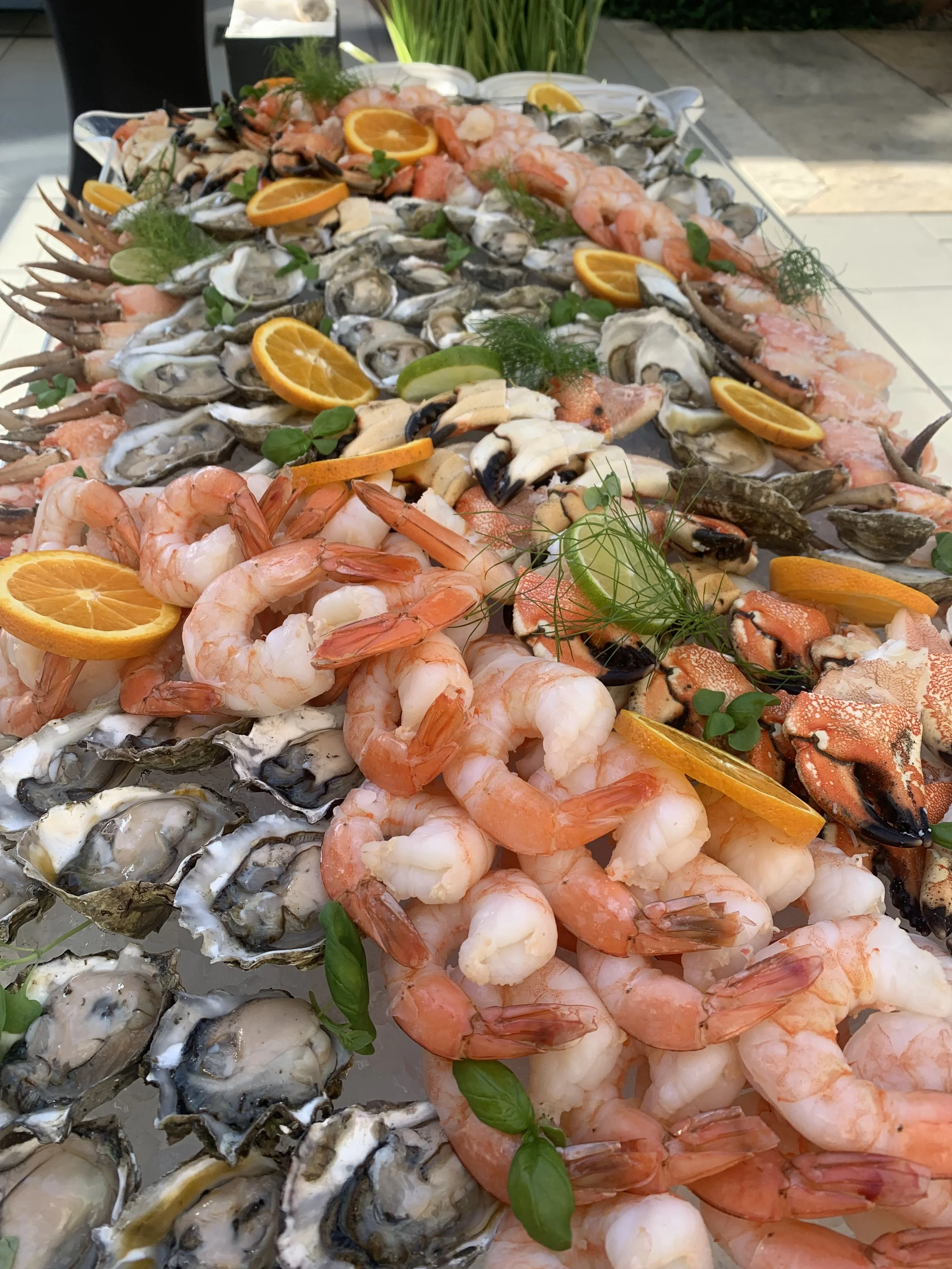 A large spread of fresh seafood, including oysters on the half shell, shrimp, crab legs, and fish, garnished with lemon and lime slices, sprigs of herbs, and greens.