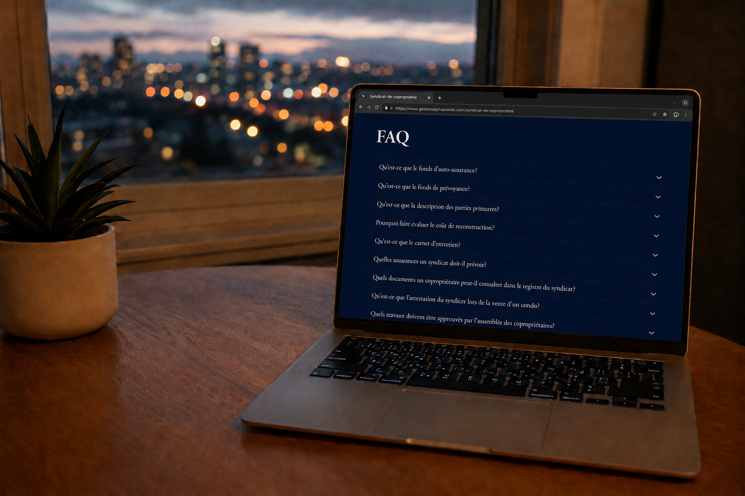 FAQ gestion copropriété - Un ordinateur portable affichant une page FAQ en français, posé sur une table en bois, avec une plante à gauche et un grand fenêtre donnant sur une ville éclatée de lumières la nuit.