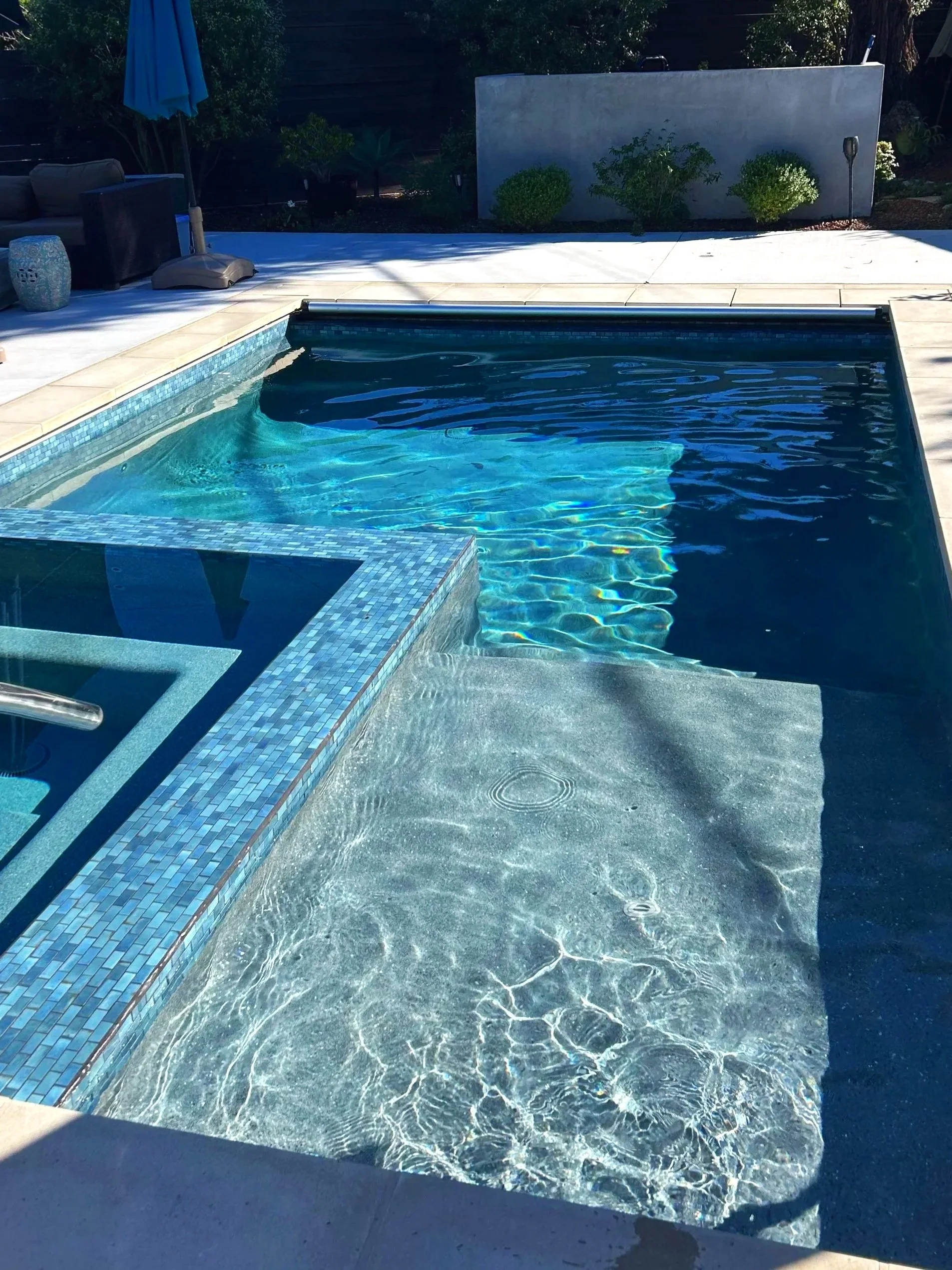 Backyard swimming pool with a waterfall feature, surrounded by a tiled deck and some outdoor furniture, plants, and a privacy wall.