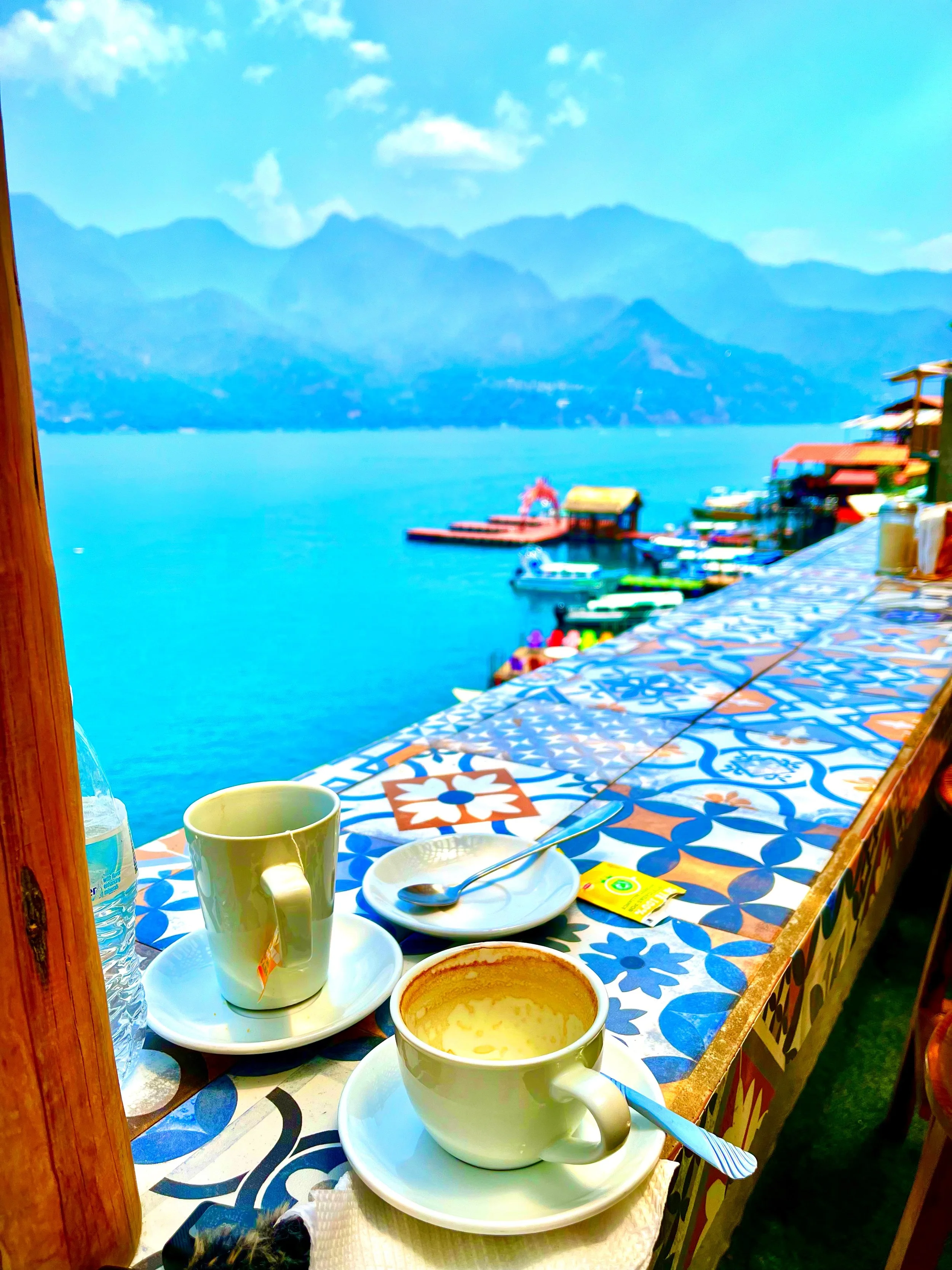 A table with coffee cups, saucers, a spoon, a water bottle, and a tea bag, overlooking a lake with mountains in the background and colorful boats docked along the shoreline.