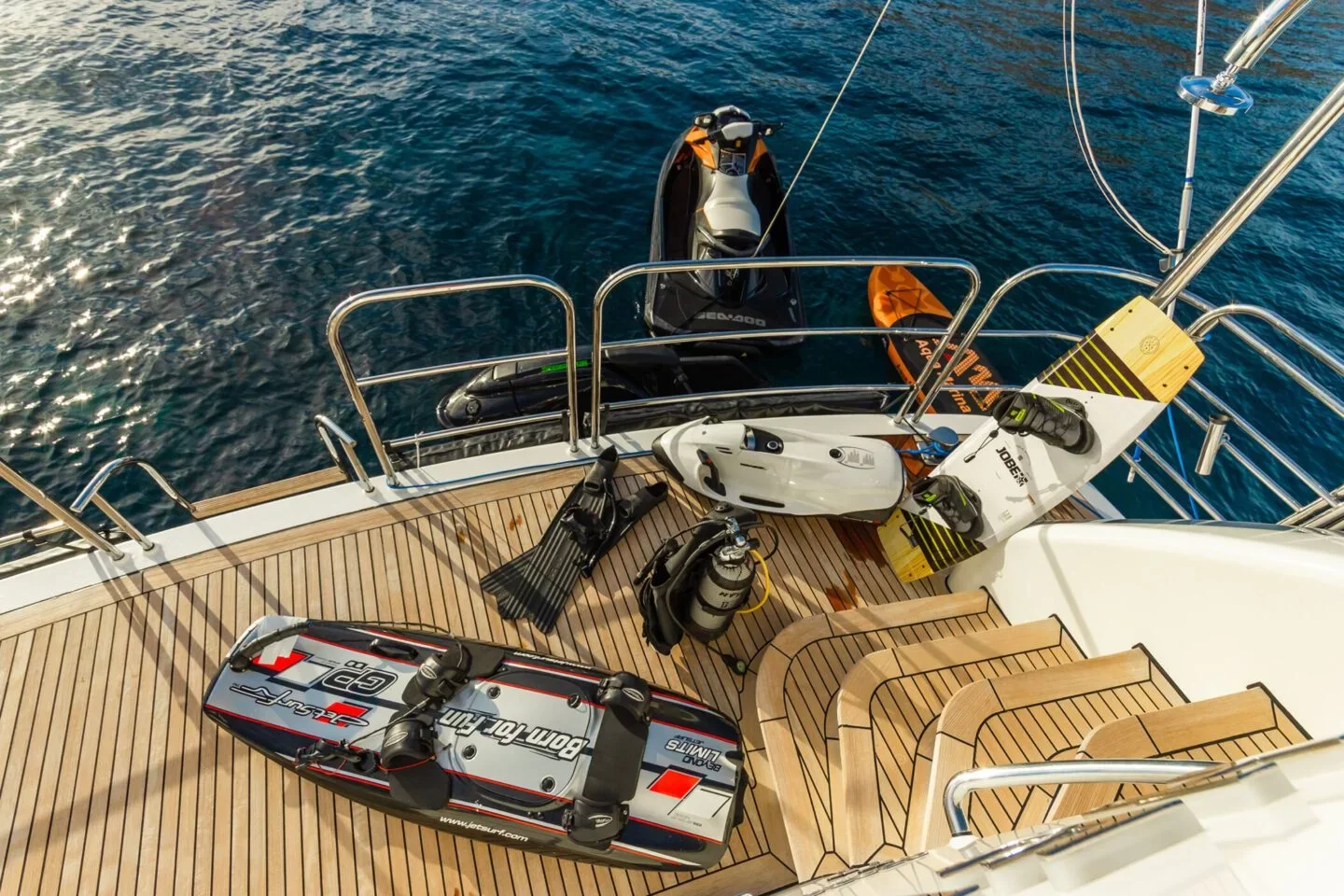 Deck of a yacht with watercraft and equipment including a jet ski, a wakeboard, a surfboard, and diving gear, with steps leading down.