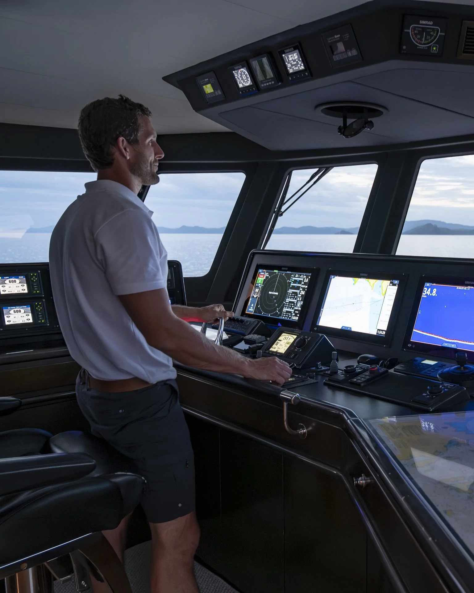A man standing at a ship's control station with multiple navigation and communication screens, looking out over the water.