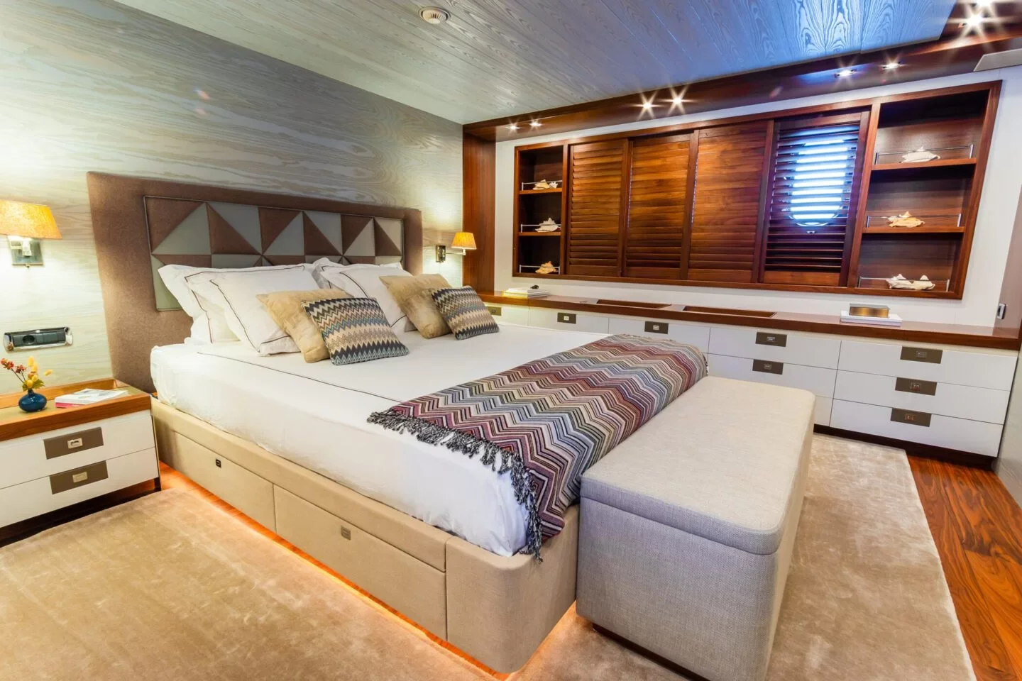 Elegant bedroom with a large bed that has decorative pillows and a colorful blanket, flanked by nightstands, with built-in wooden storage and a window with wooden shutters on the wall behind.