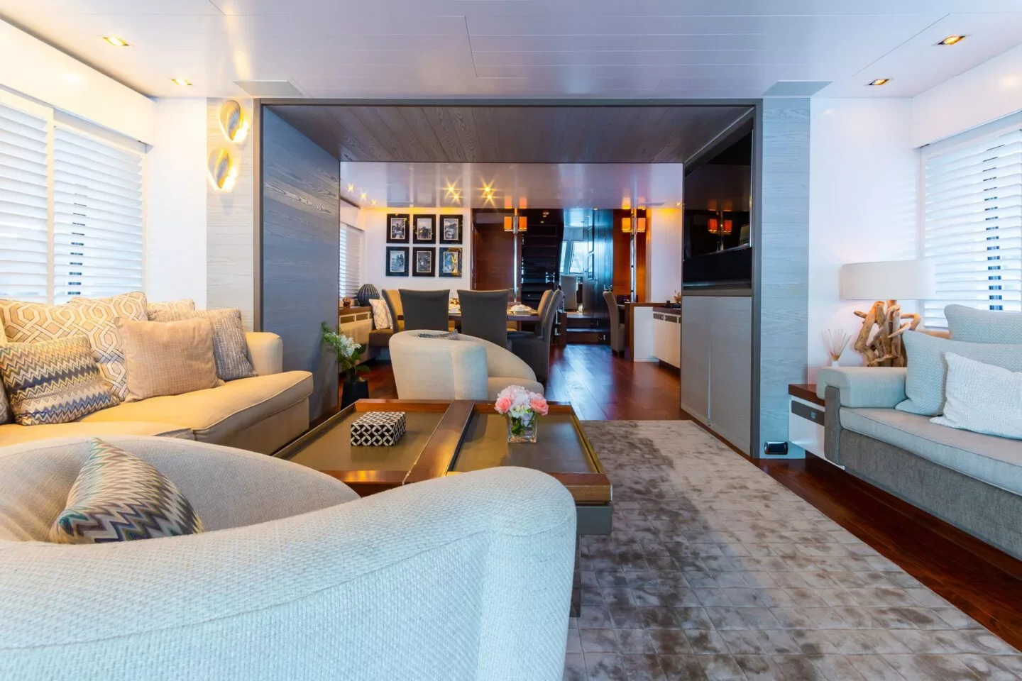 A spacious living room with light-colored sofas, patterned cushions, a wooden coffee table with a flower vase, and a large area rug on hardwood floor. The room features white shutters, modern lighting, and is open to a dining area with a table and chairs, sharing a cozy and elegant decor.