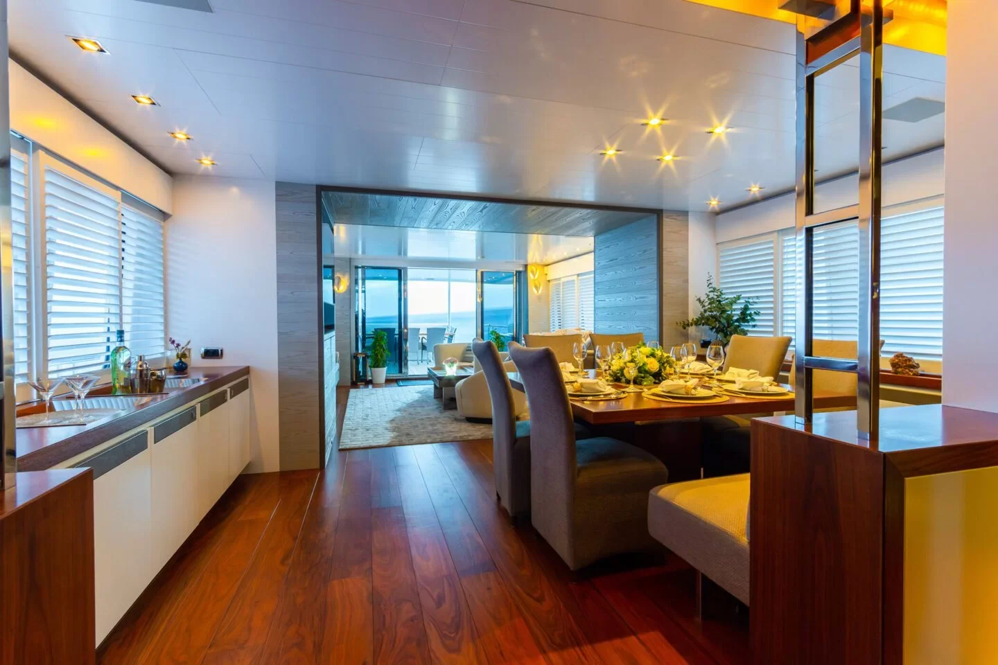 Elegant dining area with a wooden table set for a meal, surrounded by upholstered chairs, with large windows with white blinds and a view of the ocean in the background.