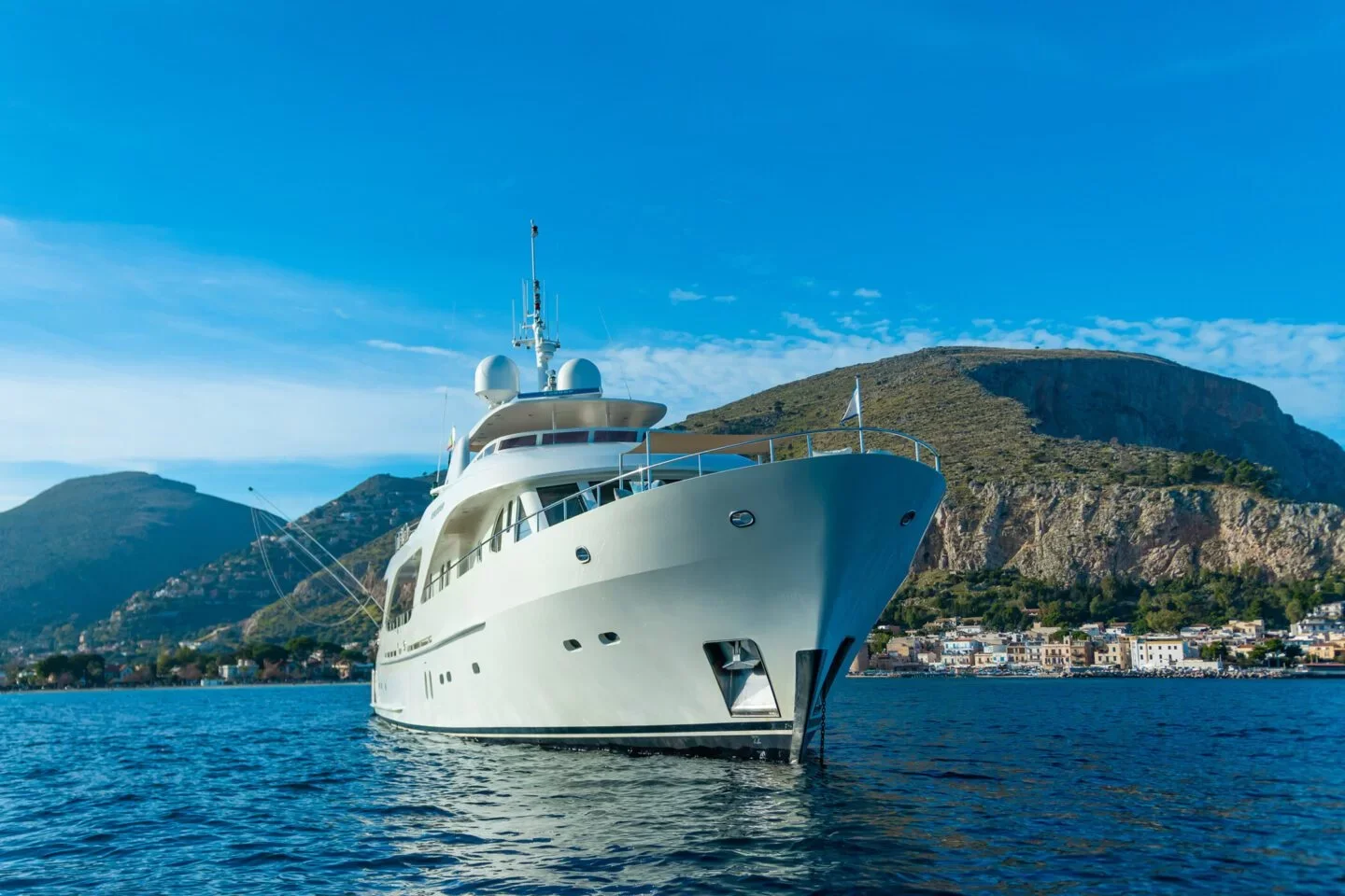 Luxury white yacht anchored in calm water with a mountain and small town in the background.