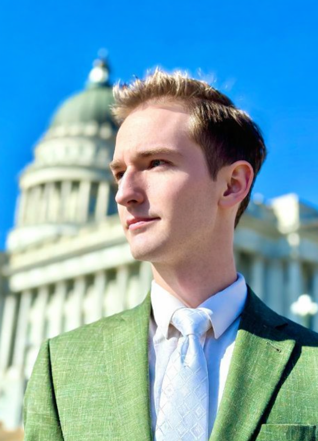 Elias Montgomery in a green suit and white tie standing outdoors in front of the Utah Capitol building on a clear, sunny day.